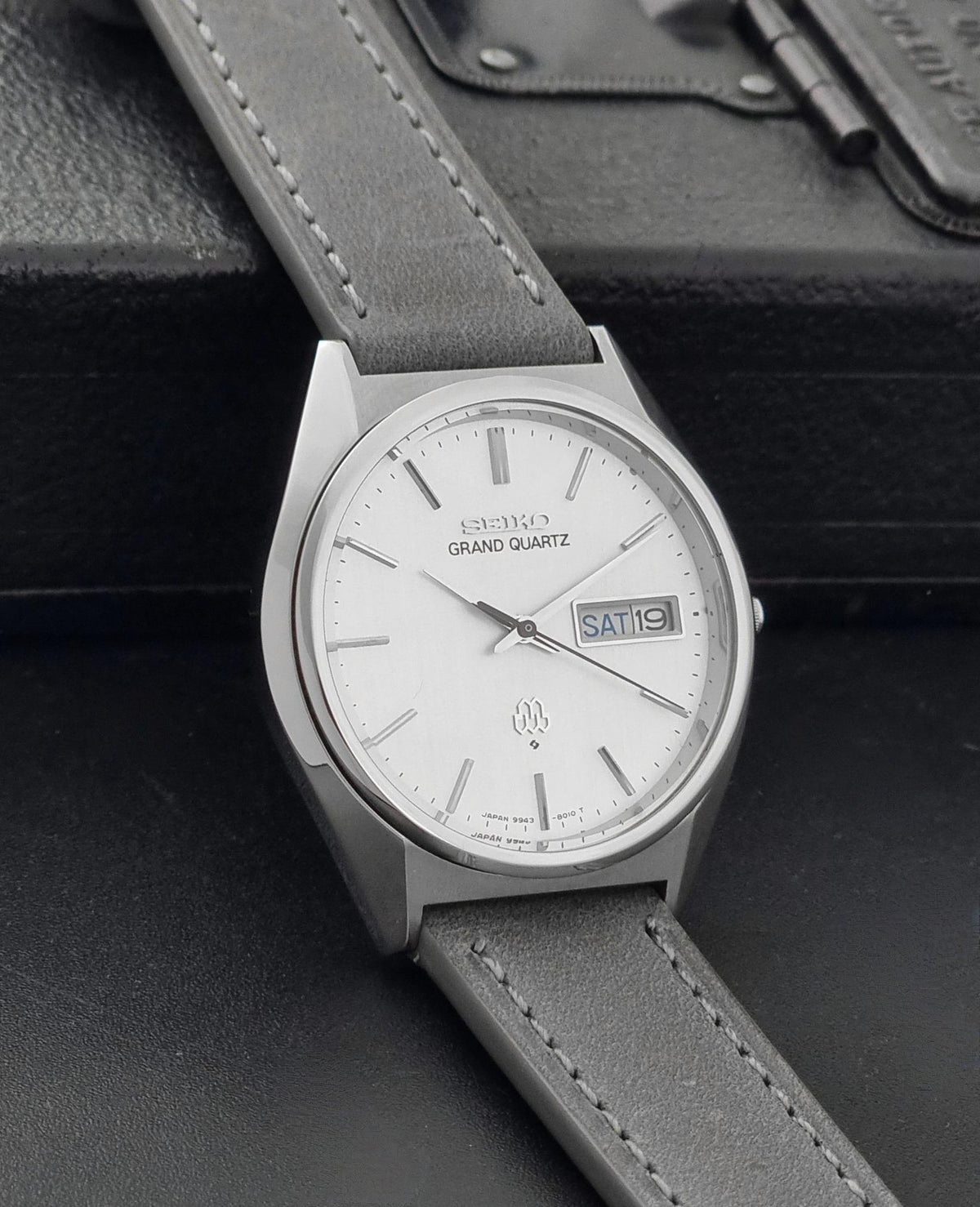 Vintage Seiko Grand Quartz 9943-8010 Wristwatch with a silver-toned metal case, white dial, and gray leather strap, resting on a dark textured surface.