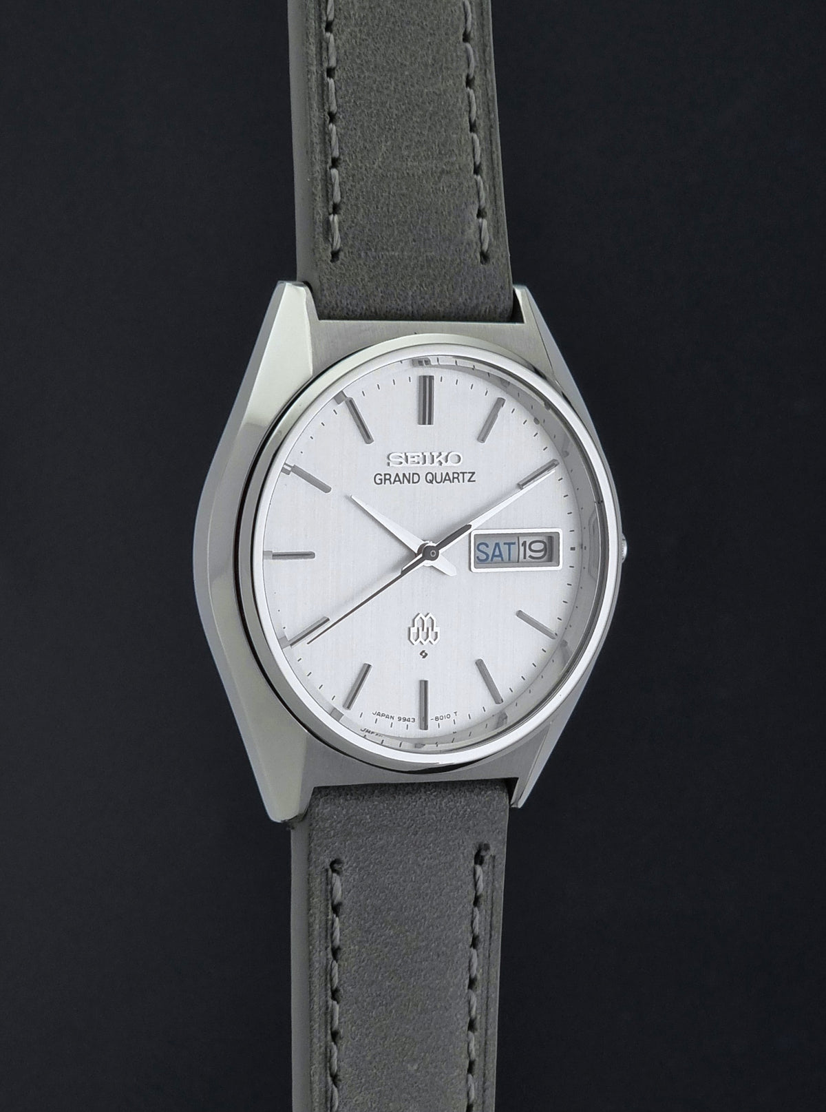 A Seiko Grand Quartz wristwatch with a silver-tone case, white dial, and a gray leather strap, shown against a dark background.