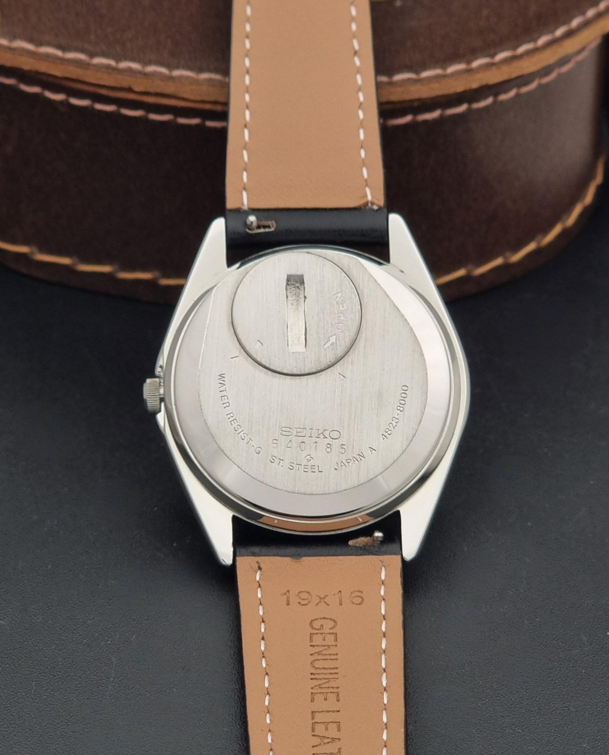 Close-up of the back of a Vintage Seiko King Quartz 4823-8000 watch with a silver metal case and a tan leather strap, resting against a dark surface with a brown leather watch box in the background.