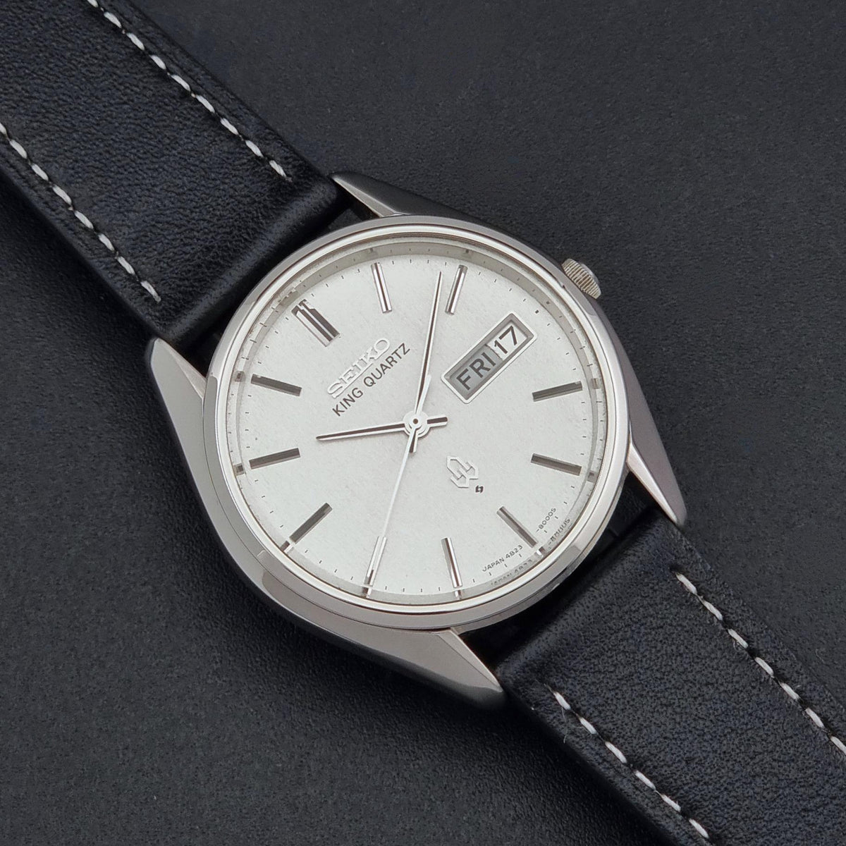 A vintage Seiko King Quartz wristwatch with a silver-toned case, white dial, and black leather strap with white stitching, shown on a dark textured background.