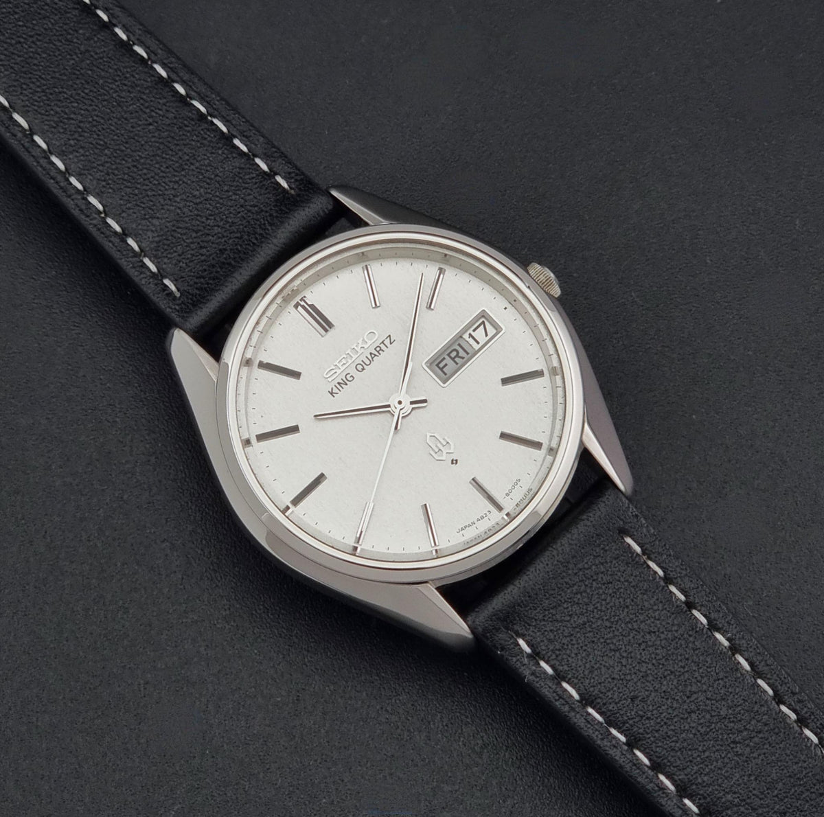 A vintage Seiko King Quartz wristwatch with a silver-toned oval case, white dial, and black leather strap with white stitching, shown diagonally on a dark textured background.