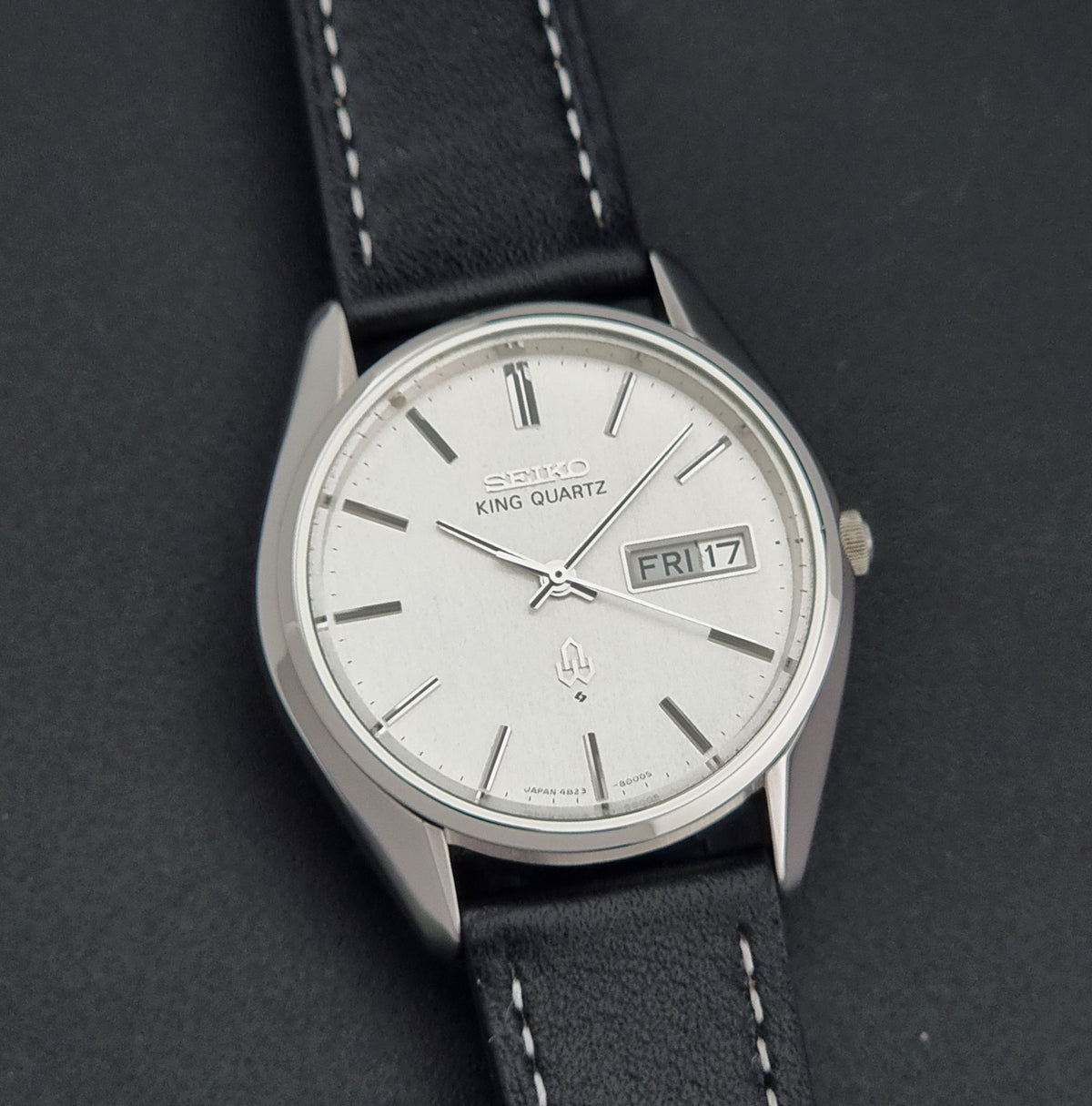 A vintage Seiko King Quartz wristwatch with a silver-toned metal case, white dial, and black leather strap with white stitching, shown on a dark textured background.
