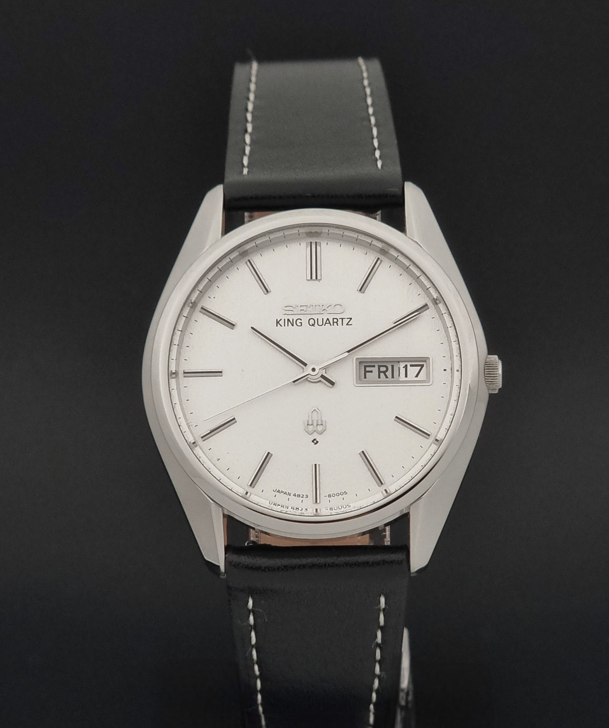 A vintage Seiko King Quartz wristwatch with a silver-toned metal case, white dial, and a black leather strap with white stitching, shown against a dark background.