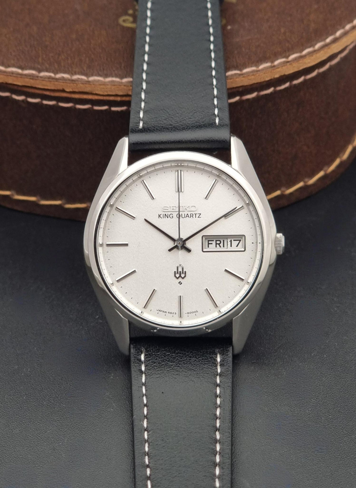 Vintage Seiko King Quartz 4823-8000 watch with a silver-toned oval case, white dial, and black leather strap with white stitching, shown resting against a brown leather surface.