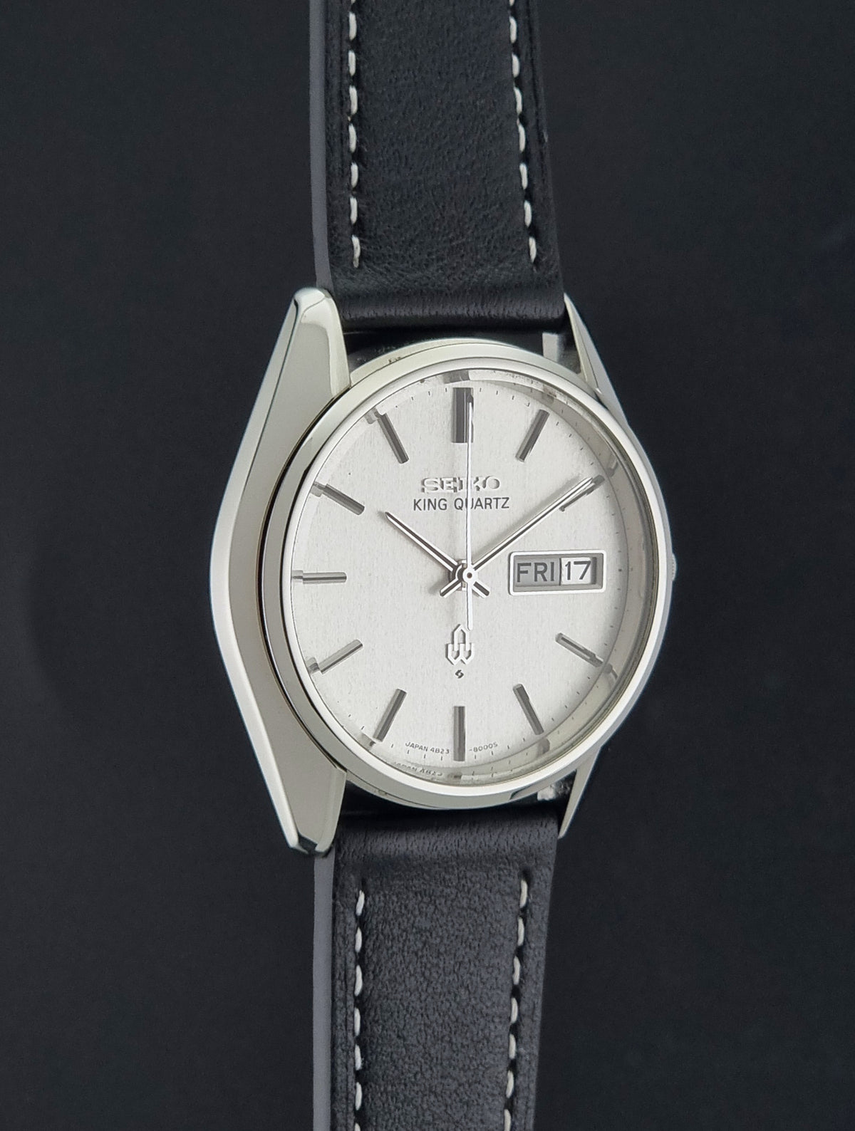 Vintage Seiko King Quartz 4823-8000 watch  with a silver-toned metal case, a white dial with stick indices and a day-date window, and a black leather strap with white stitching, shown against a dark background.