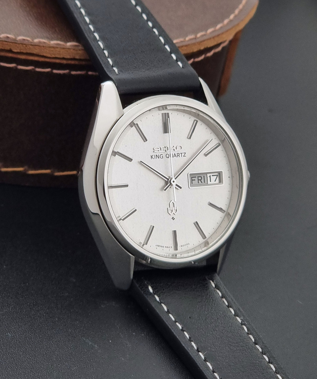 A Seiko King Quartz wristwatch with a silver-toned stainless steel case, white dial, and black leather strap with white stitching, resting on a dark surface with a brown leather case in the background.