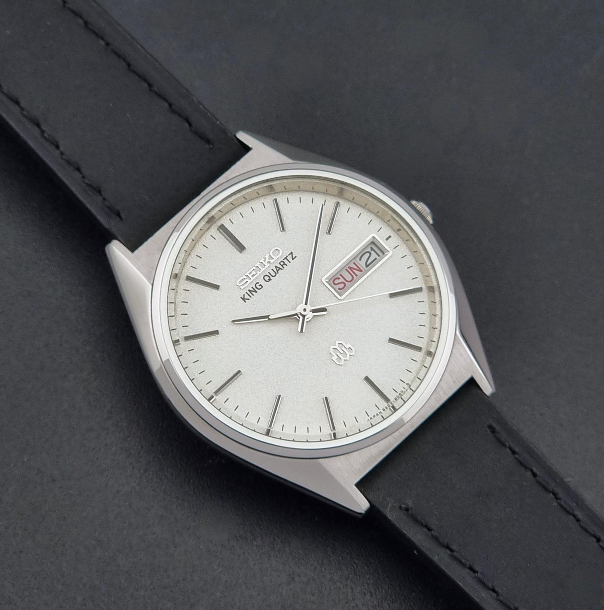 A Seiko King Quartz wristwatch with a silver-toned metal case, a white dial with stick indices, and a date window showing 'SUN 21'. It is fitted with a black leather strap and photographed diagonally on a dark textured background.