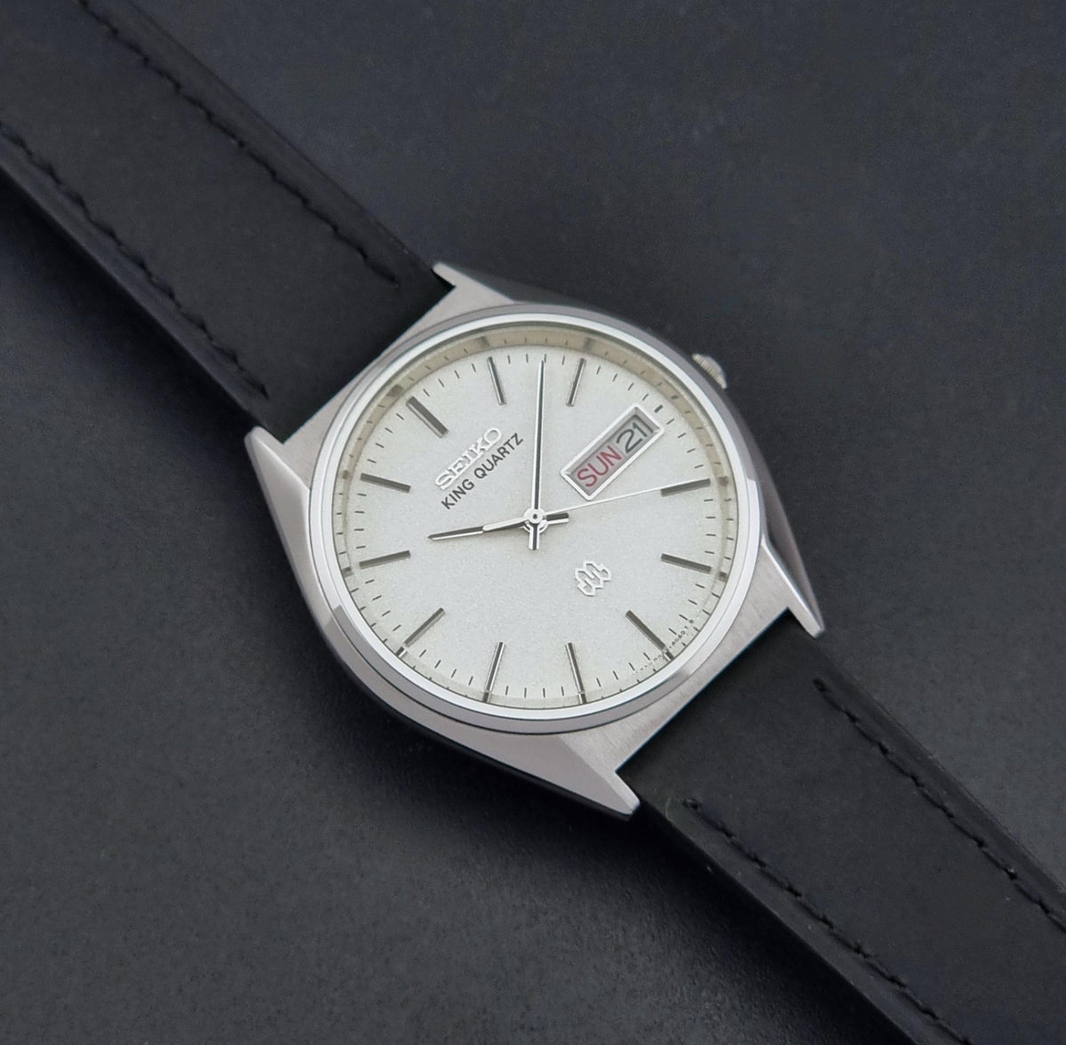 A vintage Seiko King Quartz wristwatch with a silver-toned case, white dial, and black leather strap, shown on a dark textured background.