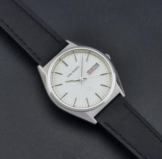 A vintage Seiko King Quartz wristwatch with a silver-toned case, white dial, and black leather strap, shown on a dark textured background.