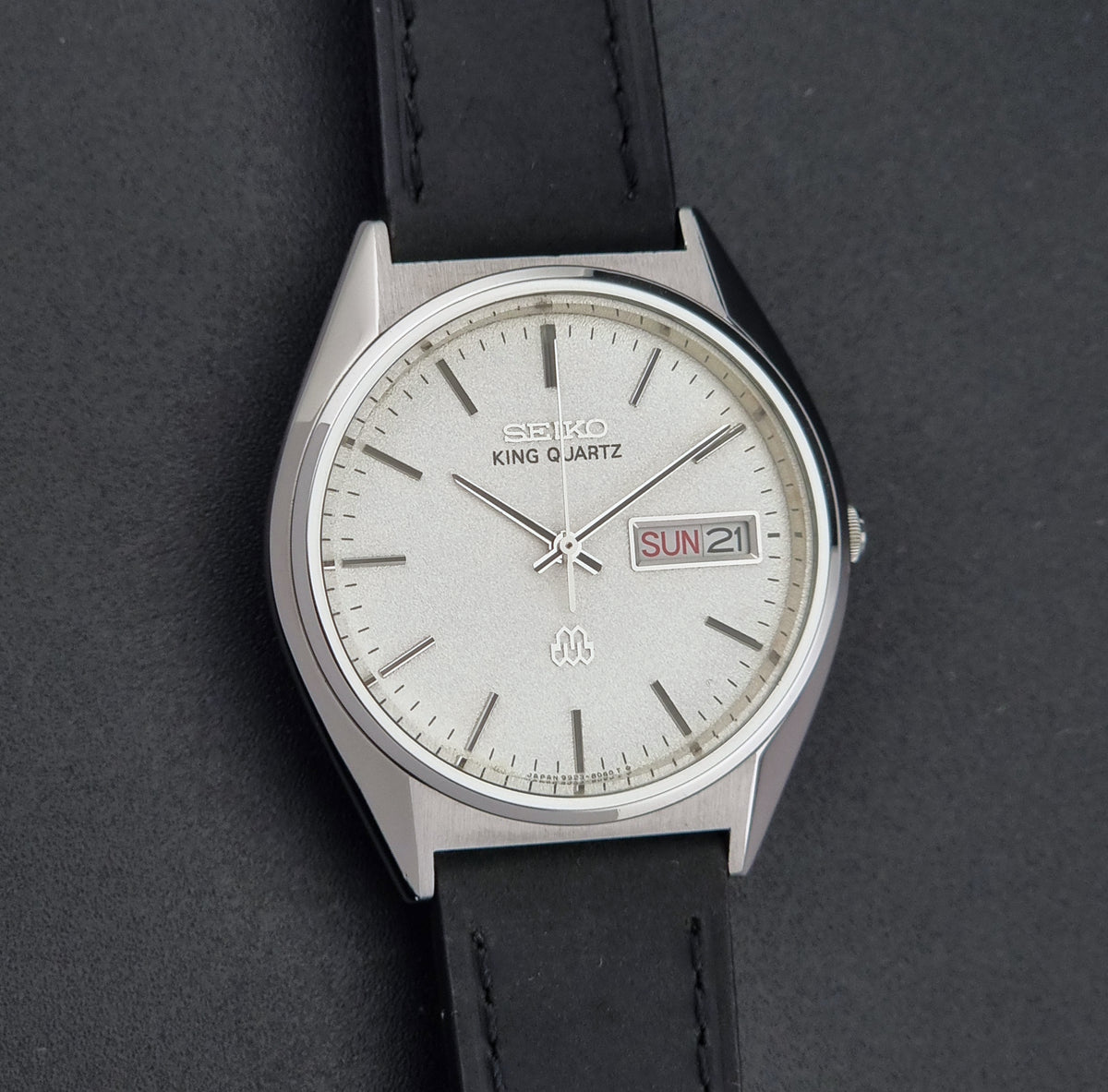 A vintage Seiko King Quartz wristwatch with a silver-toned metal case, a white dial with stick indices, and a black leather strap, shown on a dark textured background.