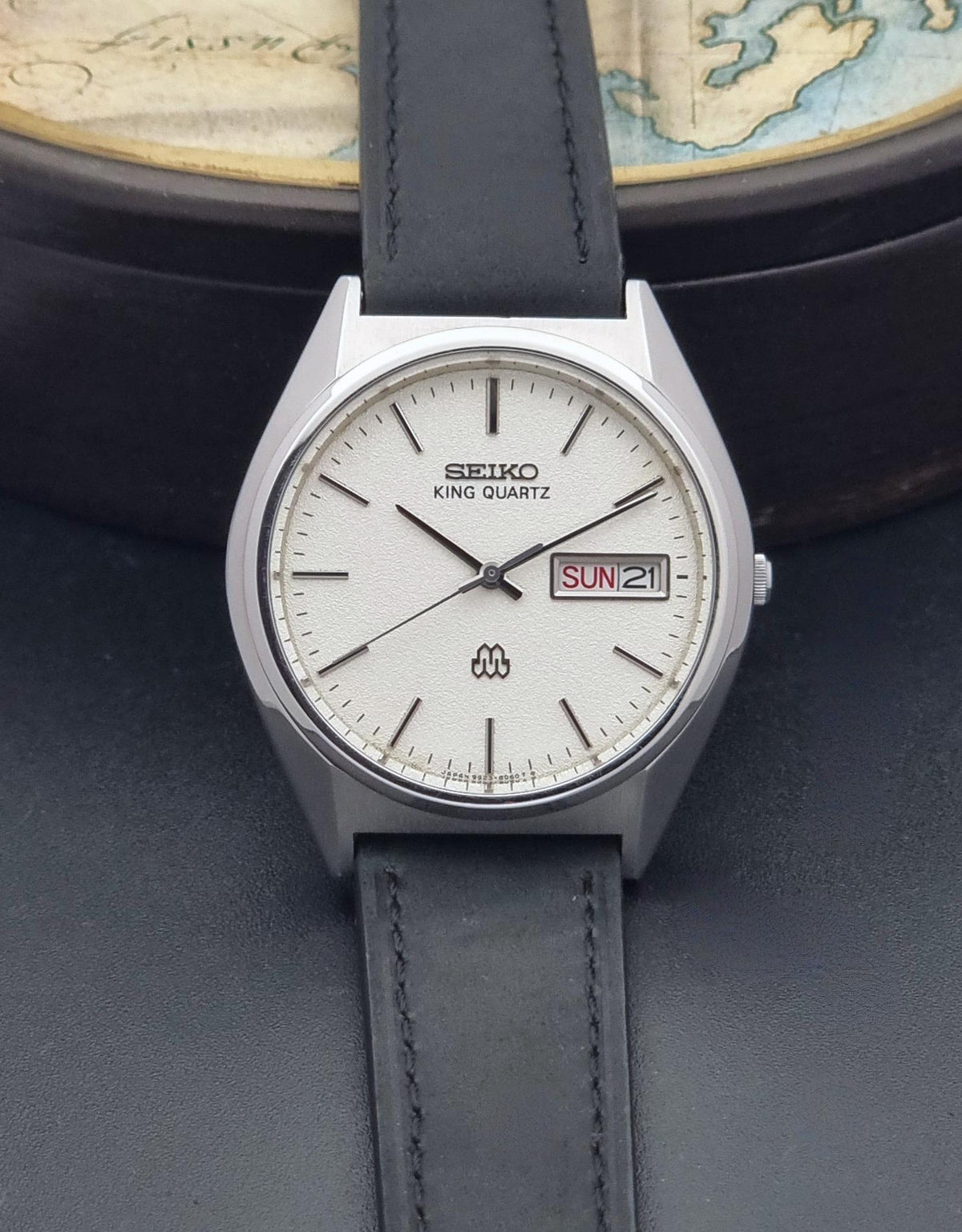 A Seiko King Quartz wristwatch with a silver-toned metal case, white dial, and black leather strap, displayed on a dark textured surface with a vintage map in the background.