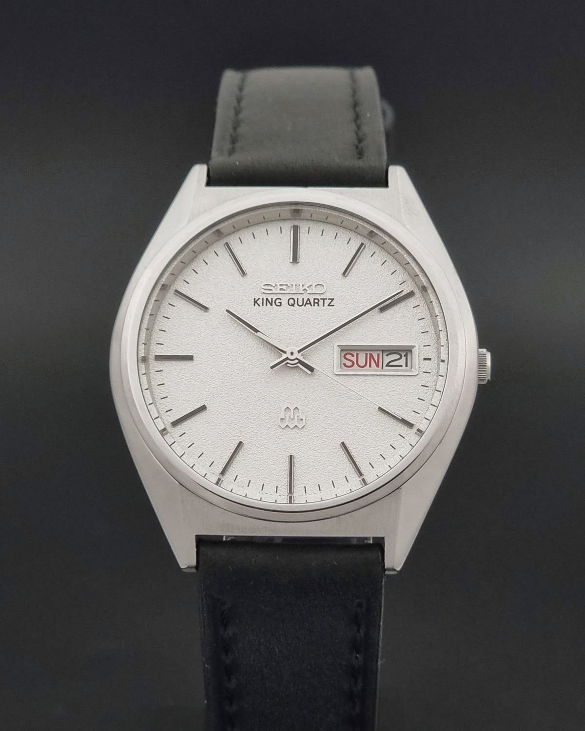 A Seiko King Quartz wristwatch with a silver-toned case, white dial, and black leather strap, resting on a dark textured surface with a vintage map box in the background.