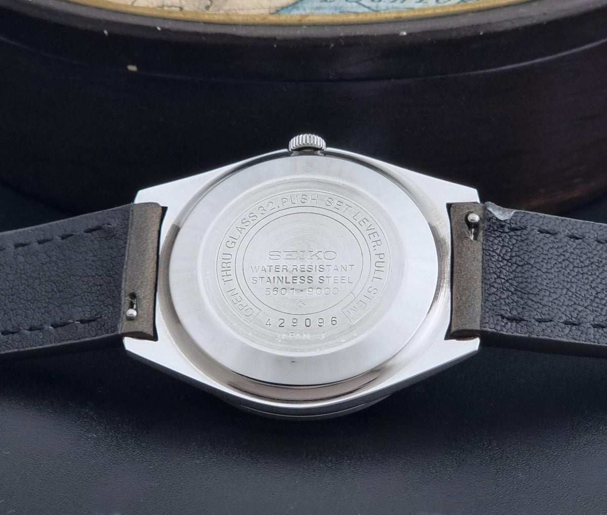Close-up of the back of a Vintage Seiko Wristwatch LordMatic 5601-9000 with a silver stainless steel case and a dark gray leather strap, resting on a dark surface.