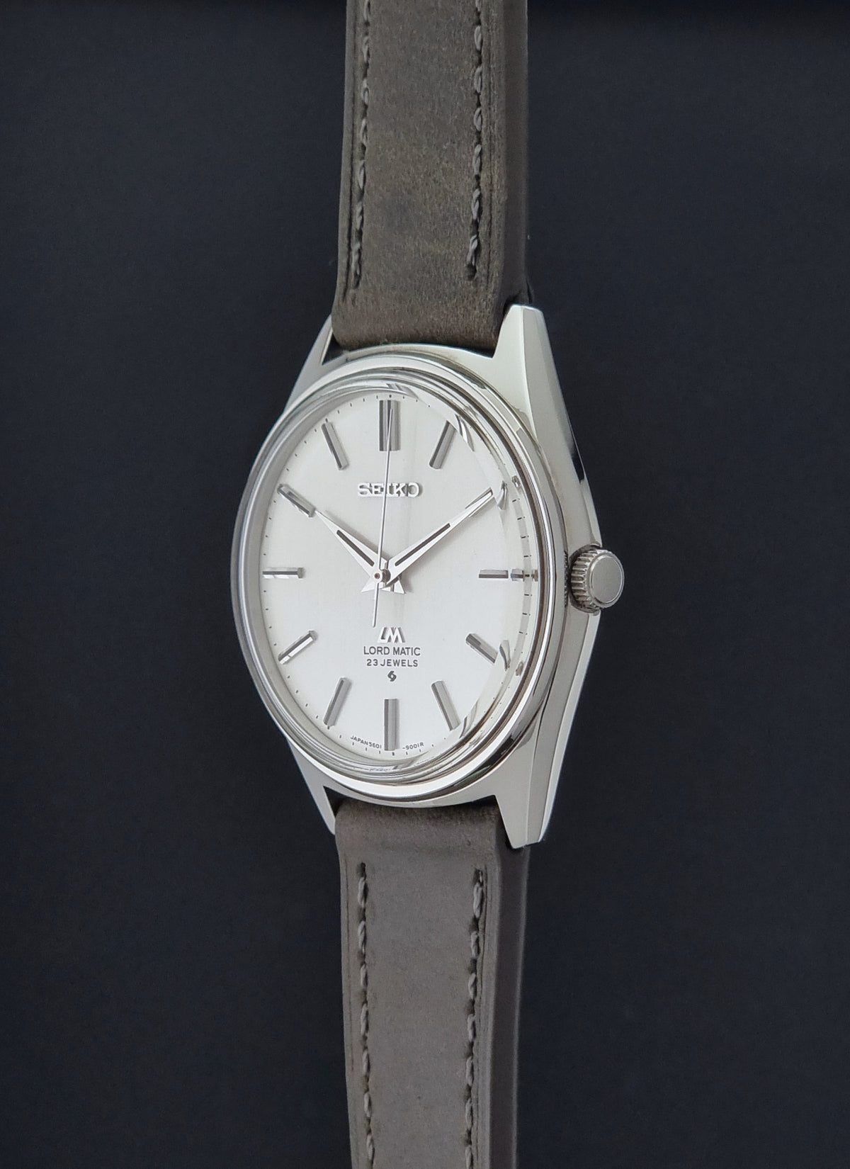 A Vintage Seiko Wristwatch LordMatic 5601-9000 with a silver-toned case, white dial, and a grey leather strap, shown against a dark background.