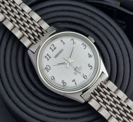 Silver Seiko Lord Matic watch with a white face on a black textured background