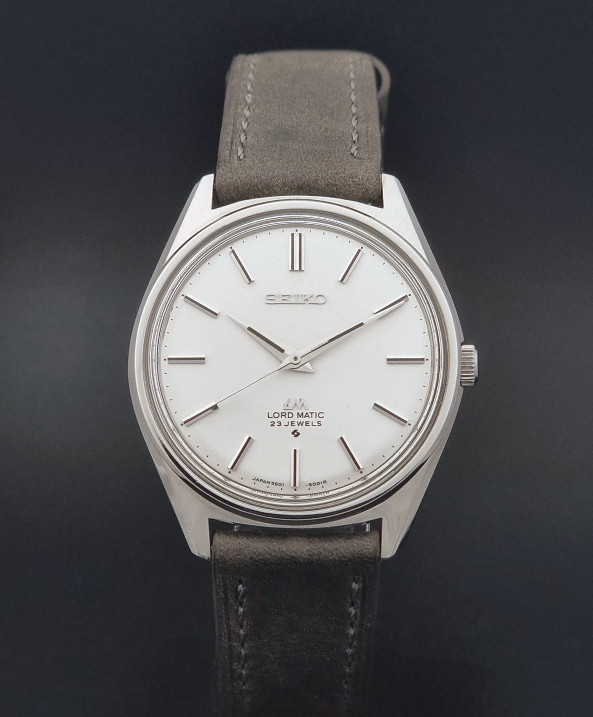 A Vintage Seiko Wristwatch LordMatic 5601-9000 with a silver-toned oval case, white dial, and a dark grey leather strap, shown against a dark background.