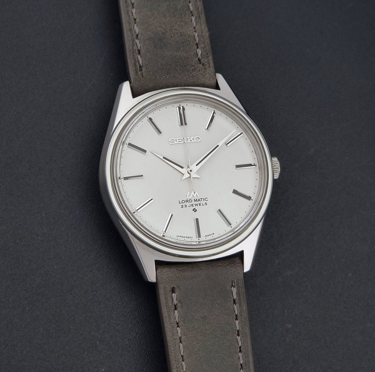 A Vintage Seiko Wristwatch LordMatic 5601-9000 with a silver-toned case, white dial, and a dark grey leather strap, shown on a dark textured background.