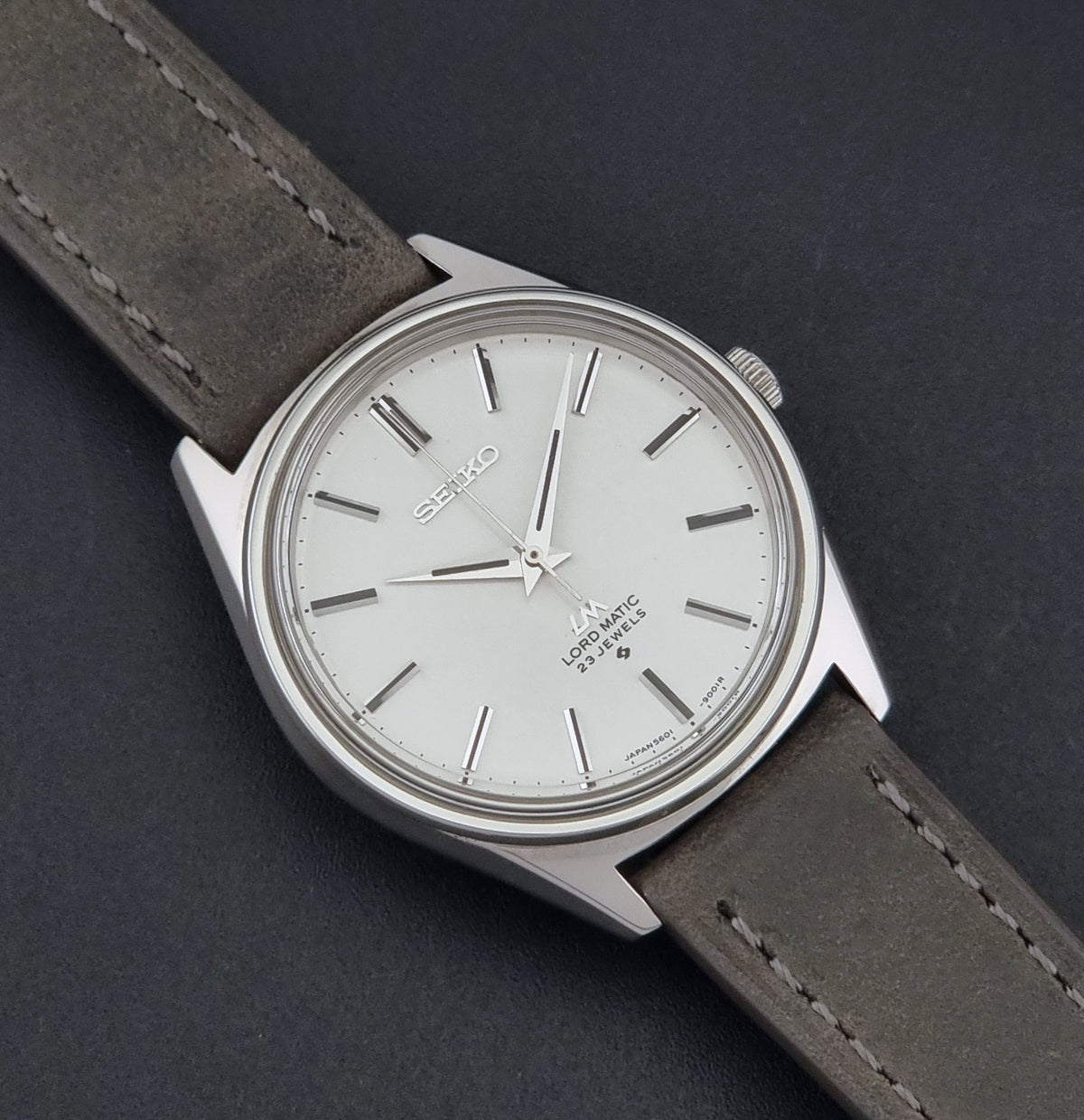 A Vintage Seiko Wristwatch LordMatic 5601-9000 with a silver-toned metal case, white dial, and a dark gray leather strap, shown diagonally on a dark textured background.