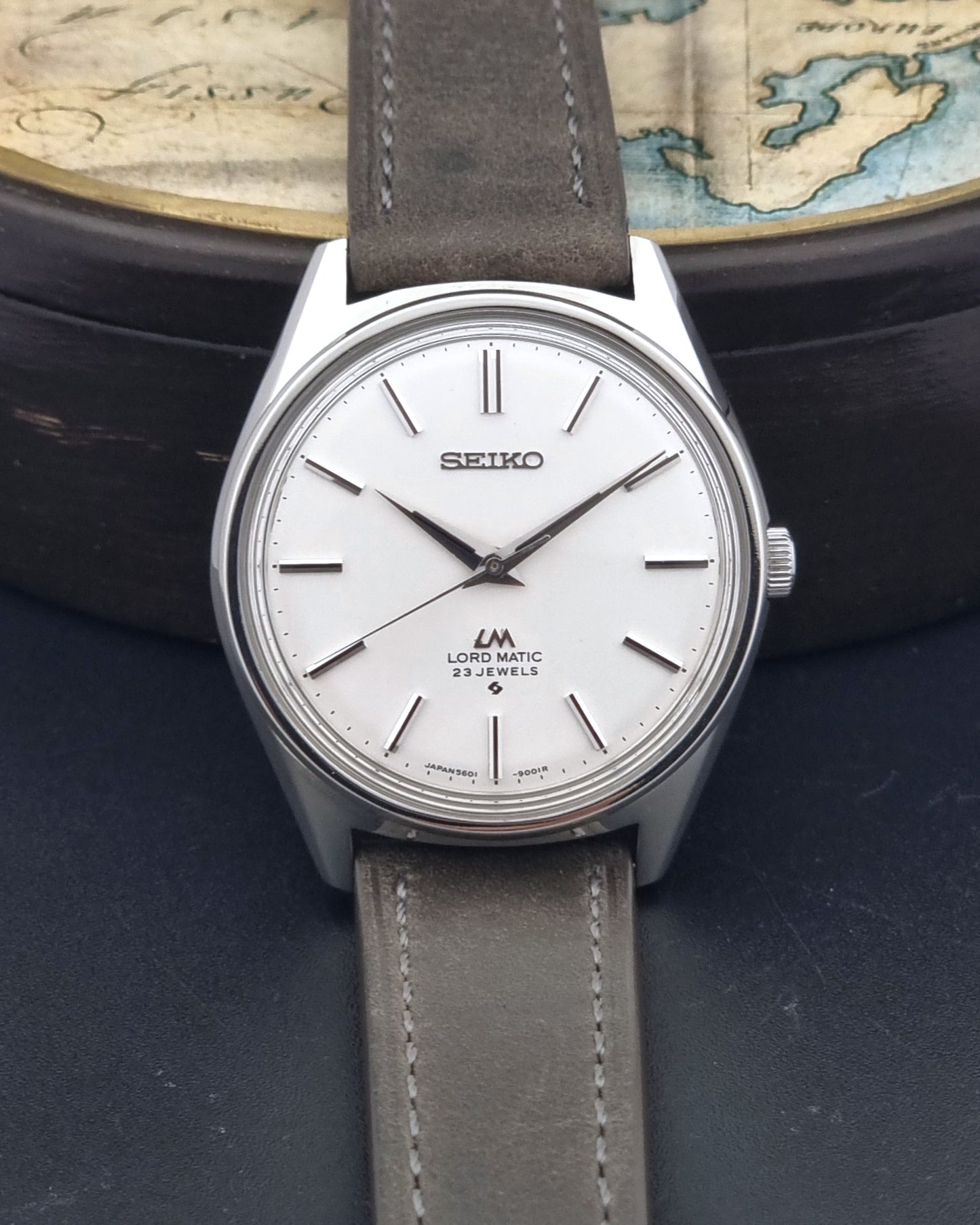 A Vintage Seiko Wristwatch LordMatic 5601-9000 with a silver-toned stainless steel case, white dial, and a grey leather strap, displayed against a dark background with a vintage map prop.