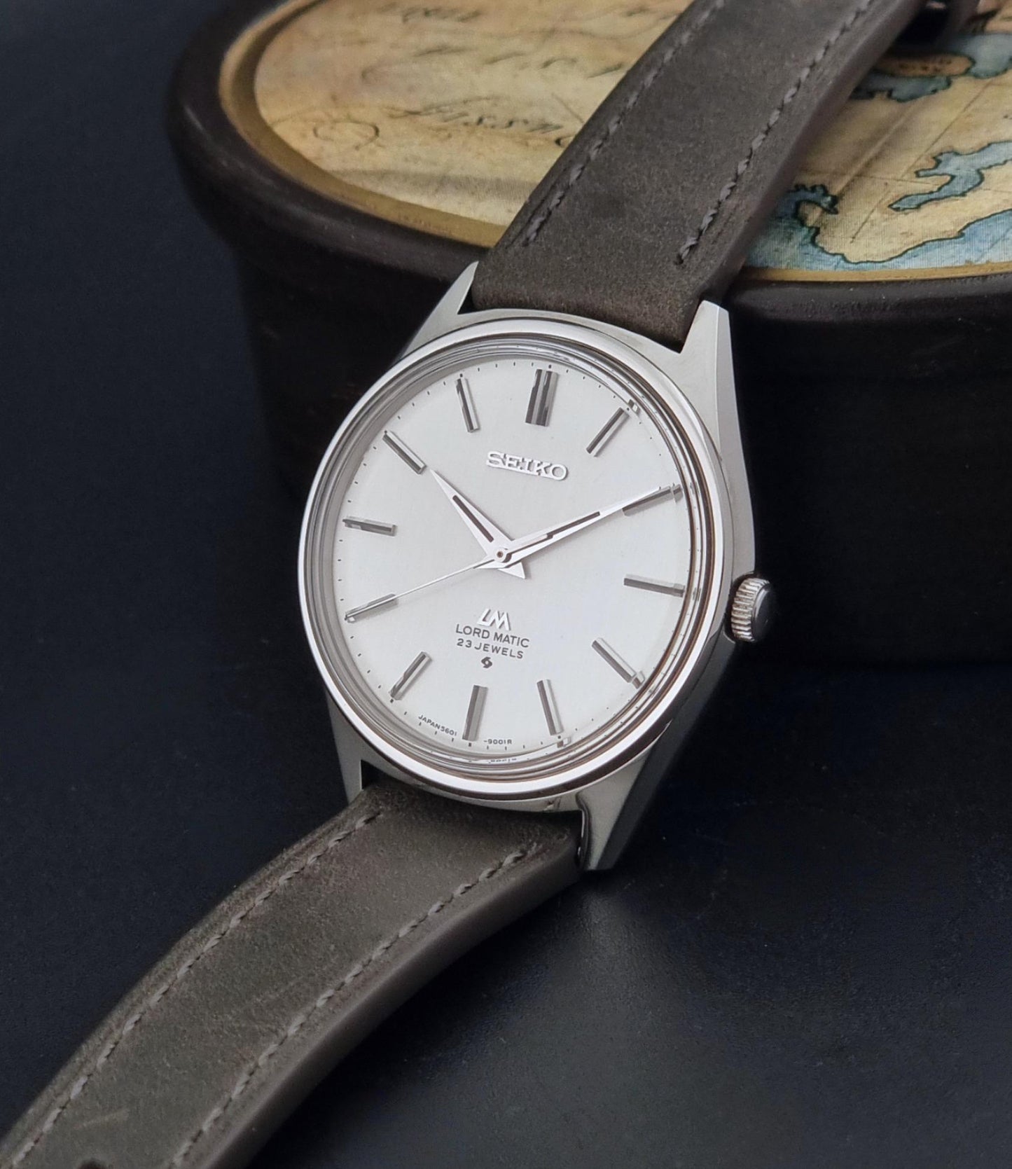 A Vintage Seiko Wristwatch LordMatic 5601-9000 with a silver-toned case, white dial, and dark gray leather strap, resting on a dark surface with a map in the background.