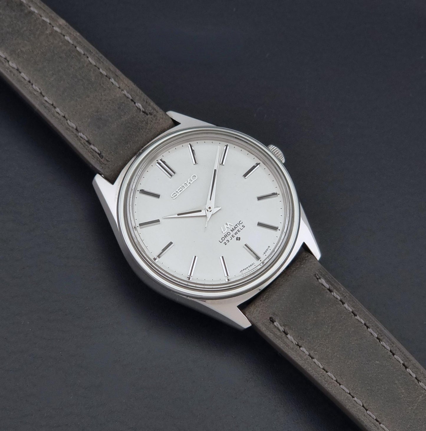 A vintage Seiko Wristwatch LordMatic 5601-9000 with a silver-toned metal case, white dial, and a dark gray leather strap, shown diagonally on a dark textured background.