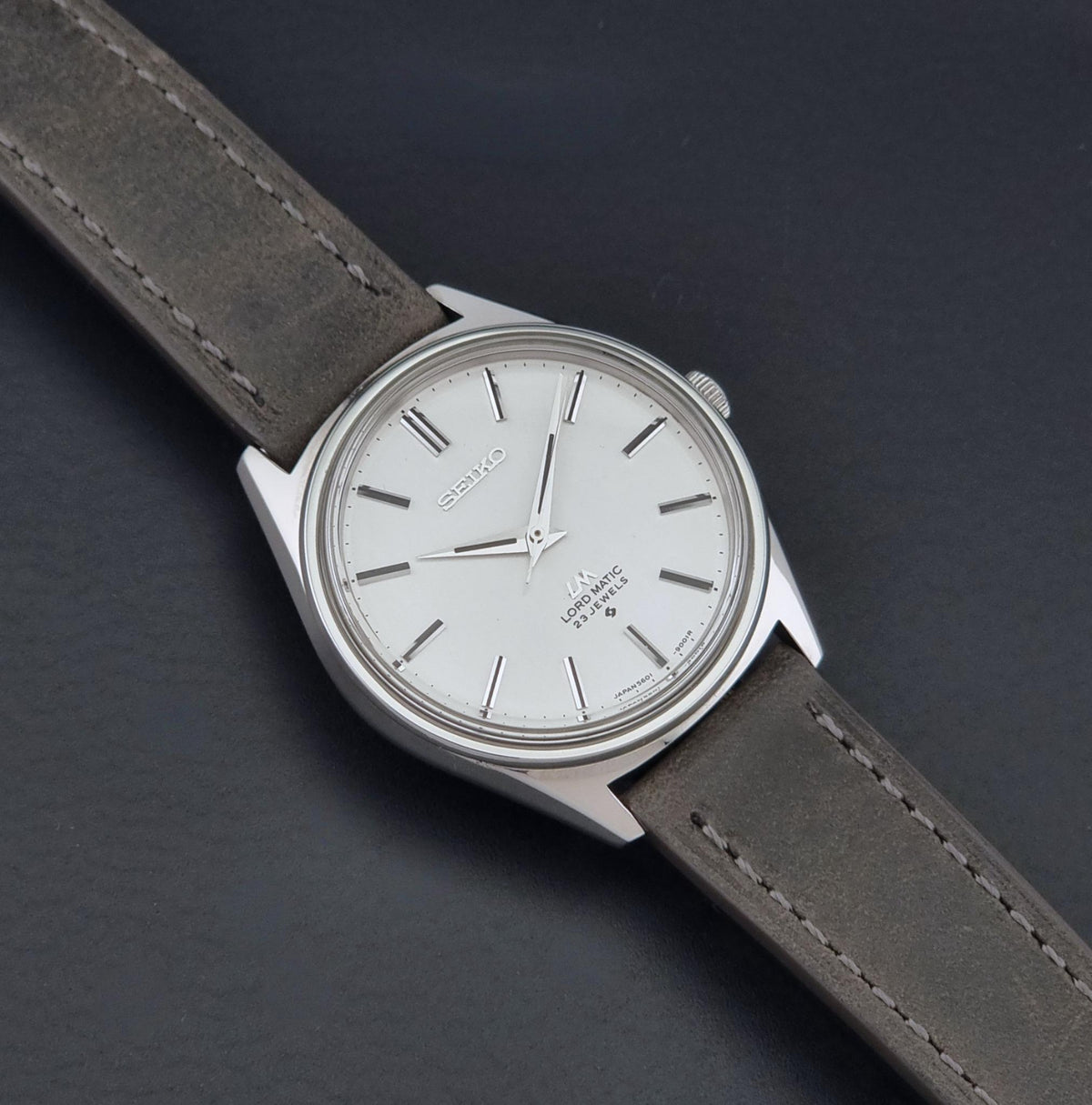 A vintage Seiko Wristwatch LordMatic 5601-9000 with a silver-toned metal case, white dial, and a dark gray leather strap, shown diagonally on a dark textured background.