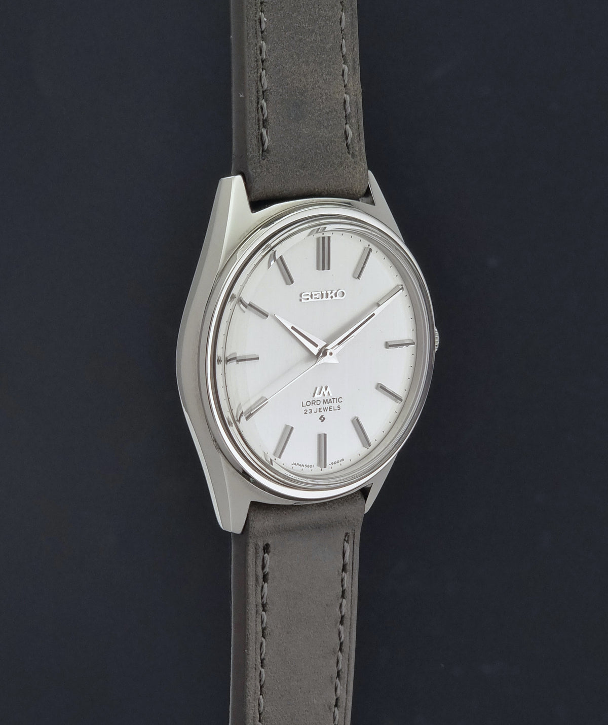 A Vintage Seiko Wristwatch LordMatic 5601-9000 with a silver-toned case, white dial, and a gray leather strap, shown against a dark background.