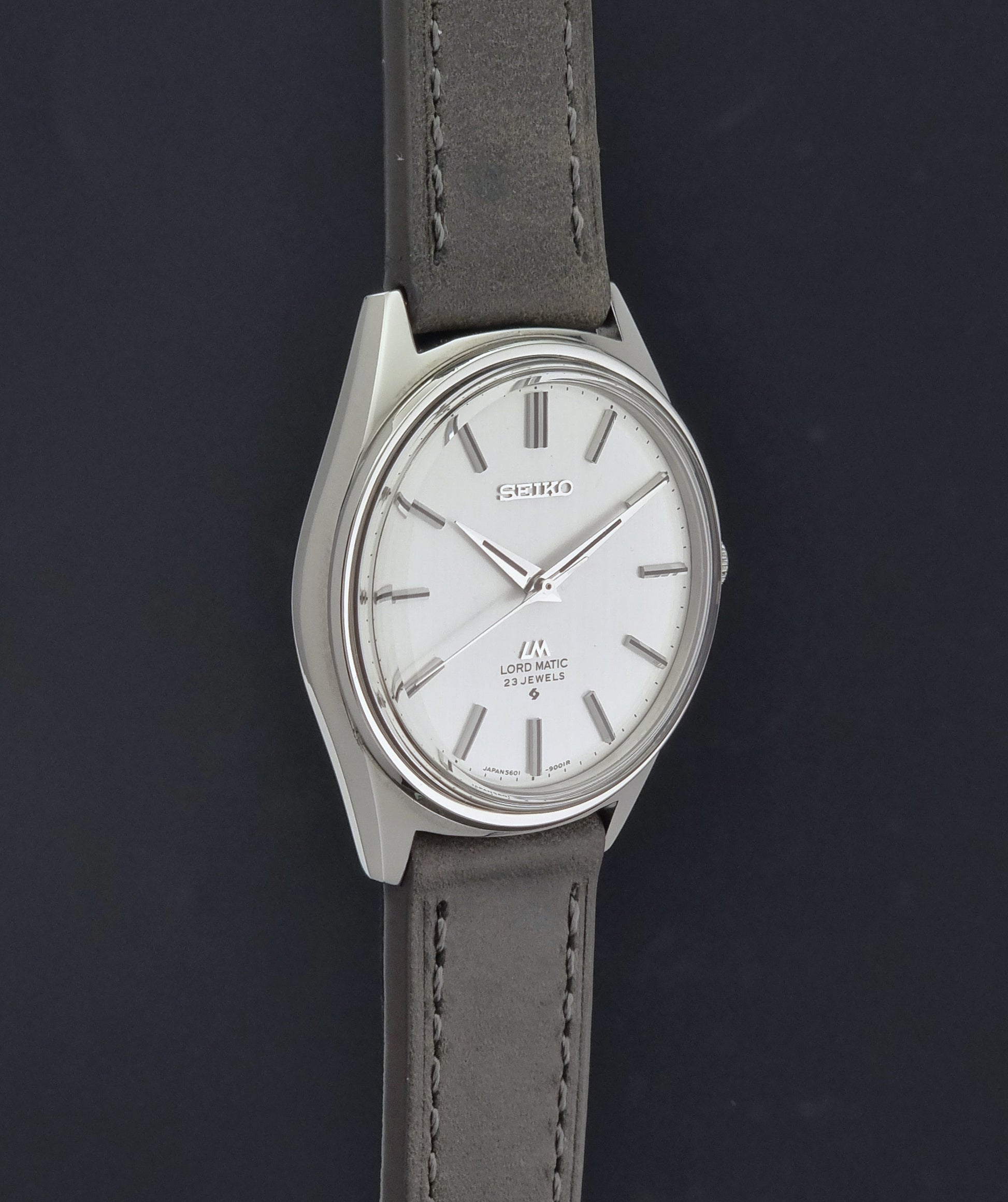 A Vintage Seiko Wristwatch LordMatic 5601-9000 with a silver-toned case, white dial, and a gray leather strap, shown against a dark background.
