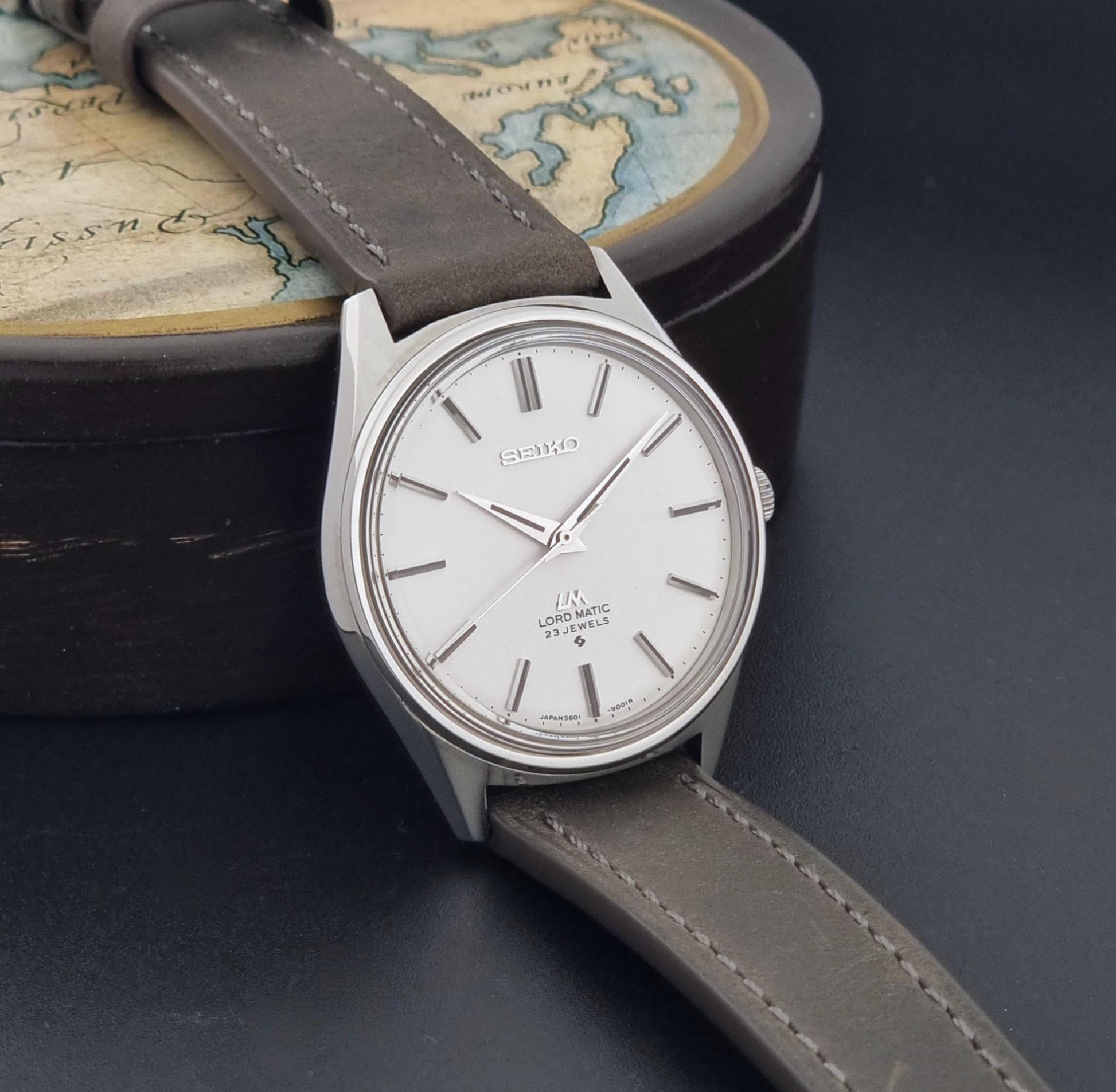 A vintage Seiko Wristwatch LordMatic 5601-9000 with a silver-toned case, white dial, and grey leather strap, resting on a dark surface with a round map box in the background.