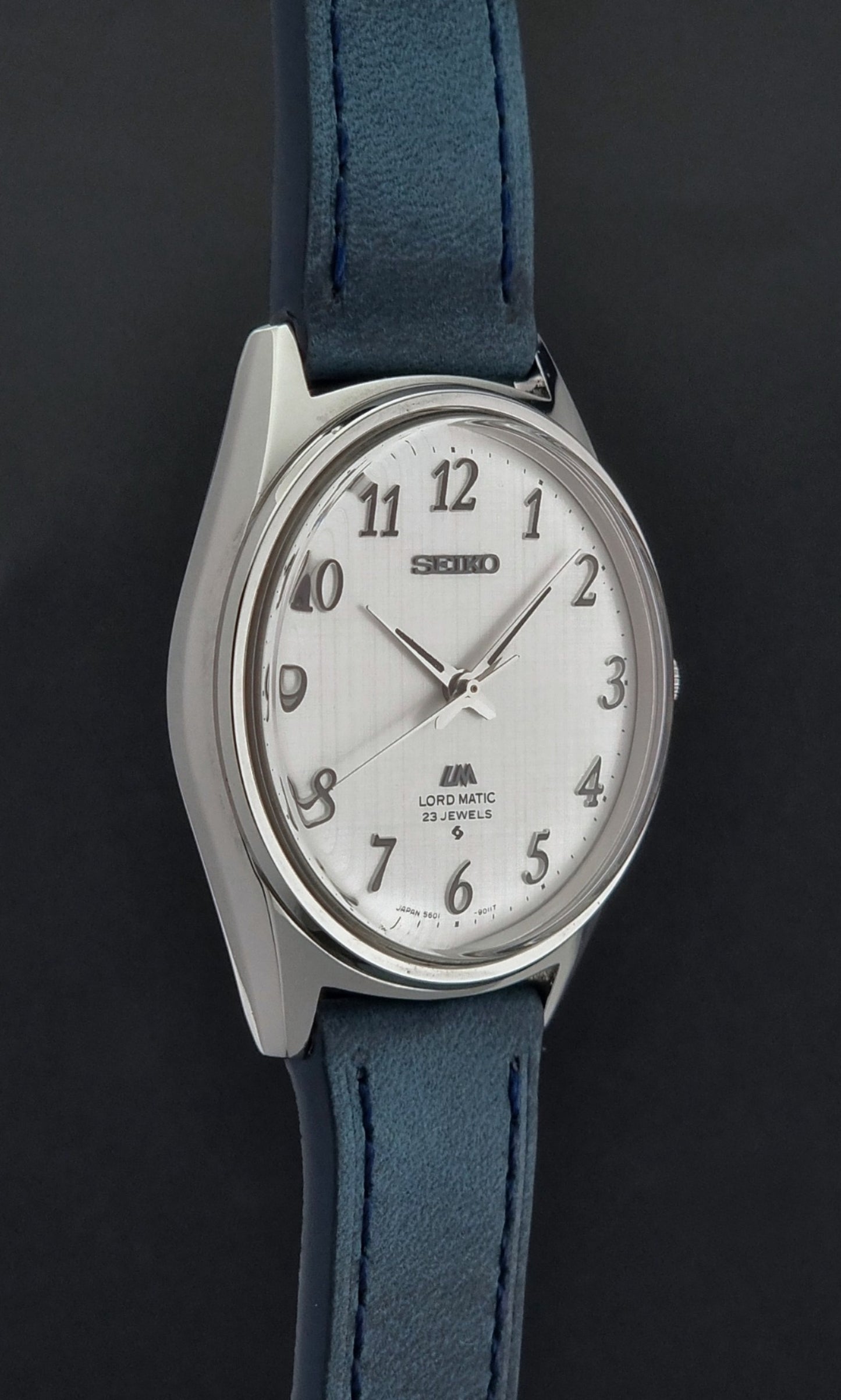 Vintage Seiko Wristwatch LordMatic 5601-9000 with blue strap and silver case on a gray background side profile view