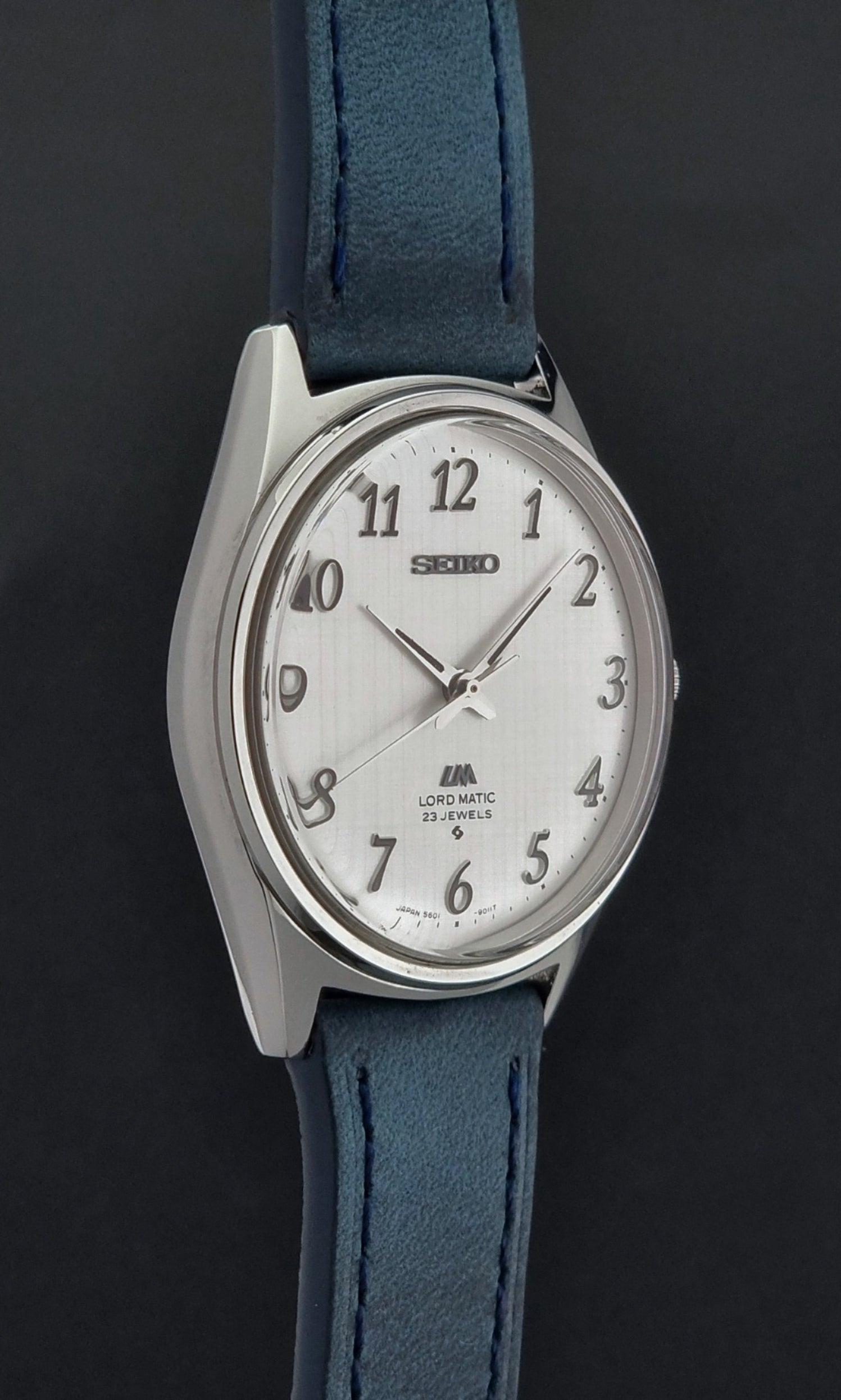 Vintage Seiko Wristwatch LordMatic 5601-9000 with blue strap and silver case on a gray background side profile view