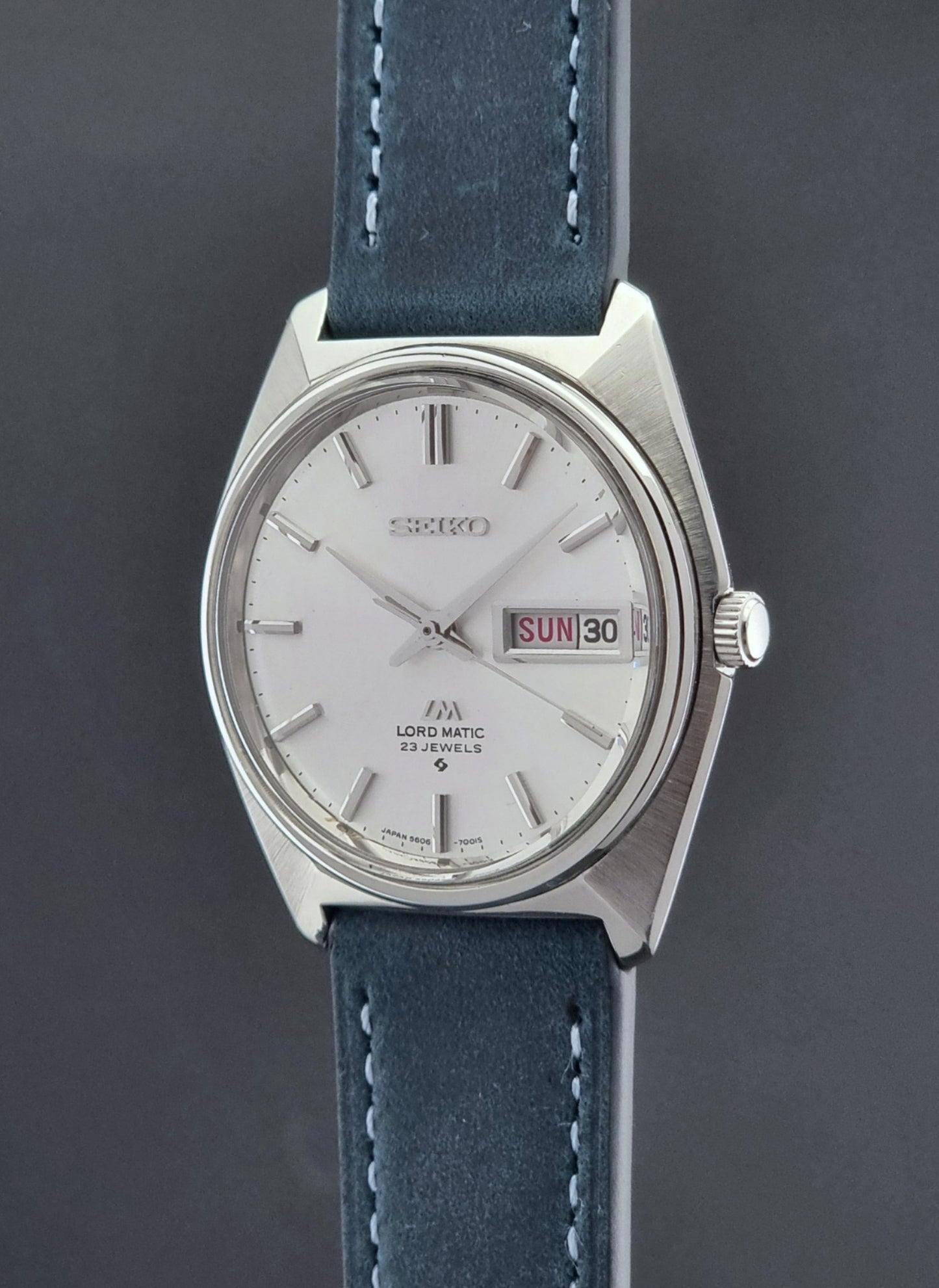 Vintage Seiko Wristwatch LordMatic 5606-7000 with blue strap on a textured gray background crown view profile