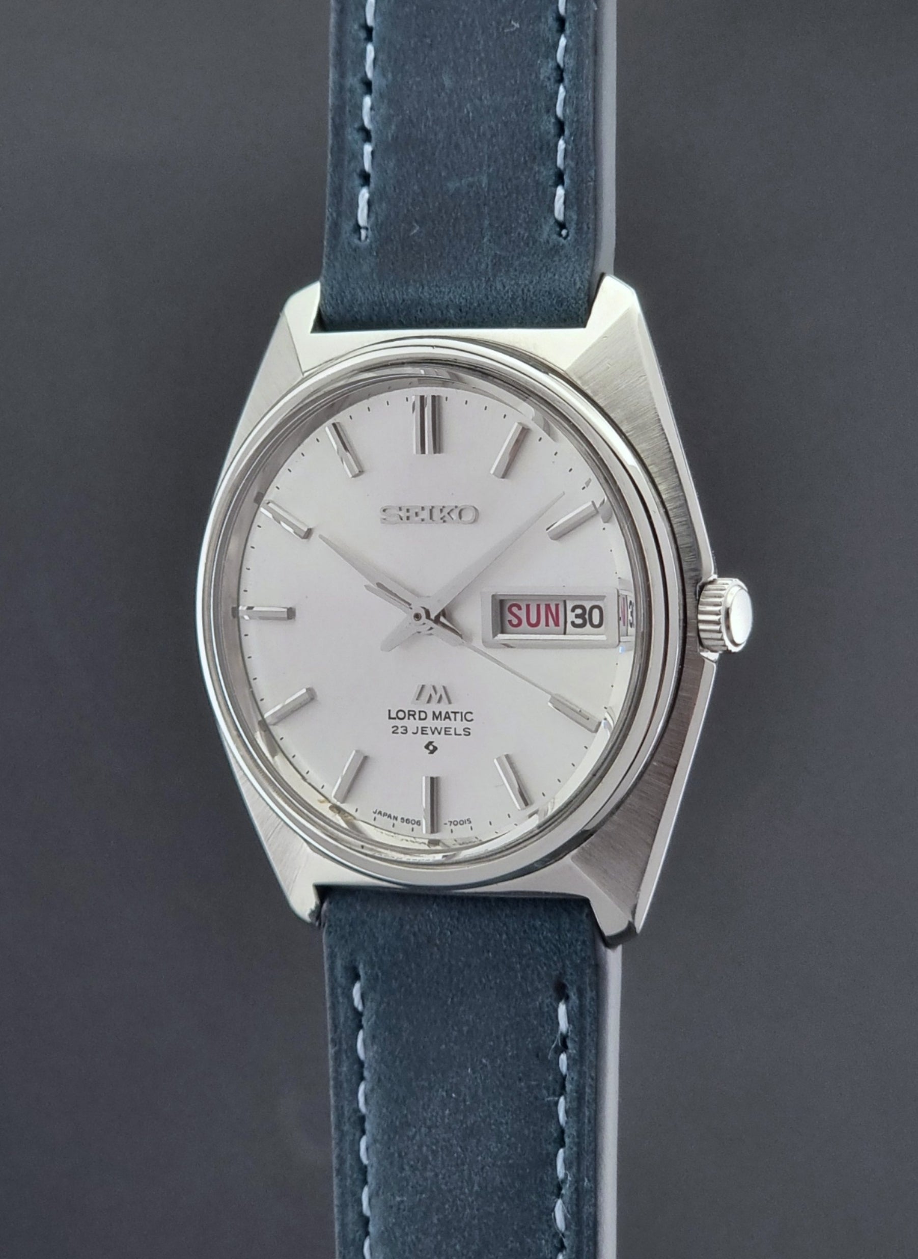Vintage Seiko Wristwatch LordMatic 5606-7000 with blue strap on a textured gray background crown view profile
