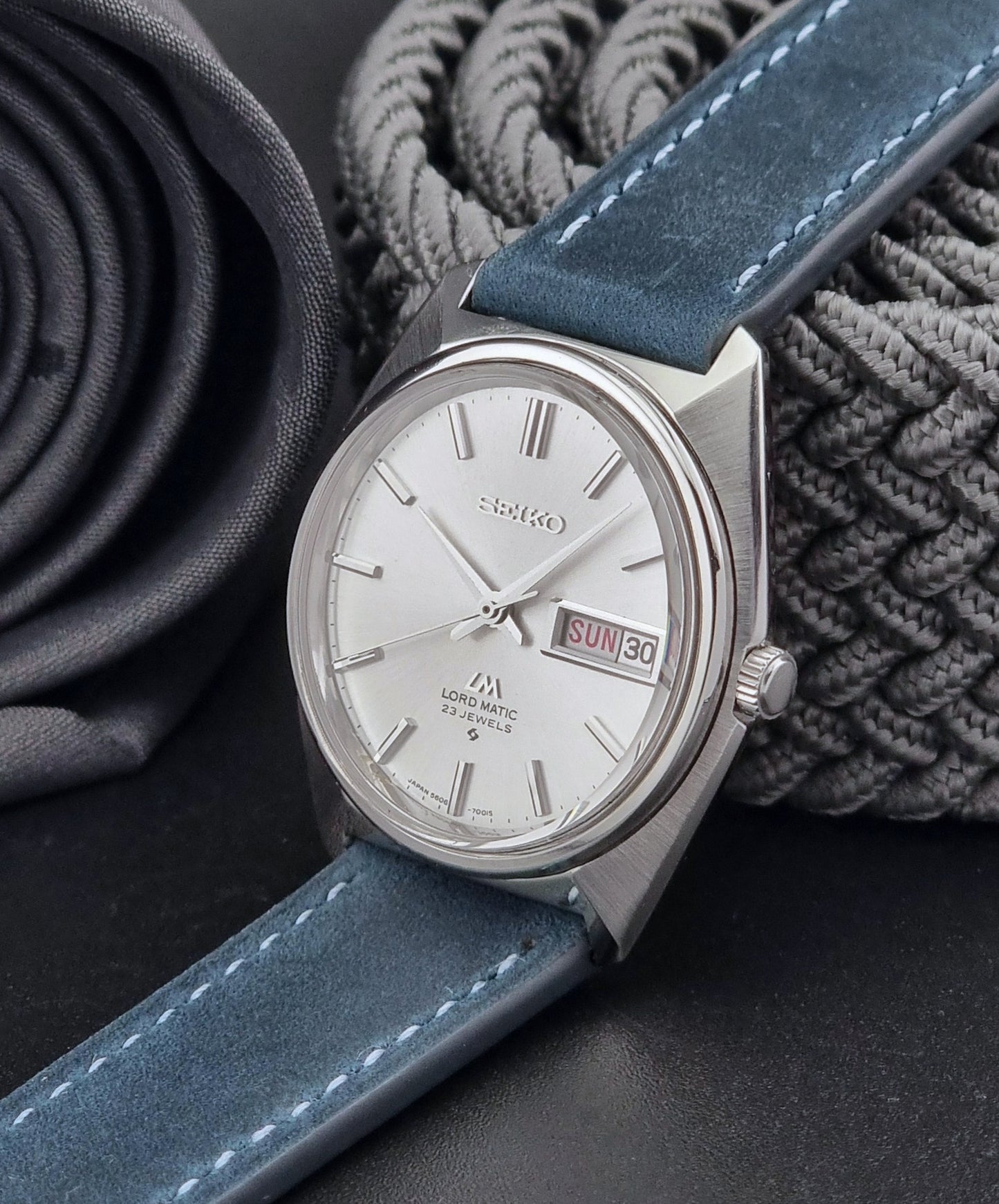 Vintage Seiko Wristwatch LordMatic 5606-7000 with blue strap on a textured gray background crown view