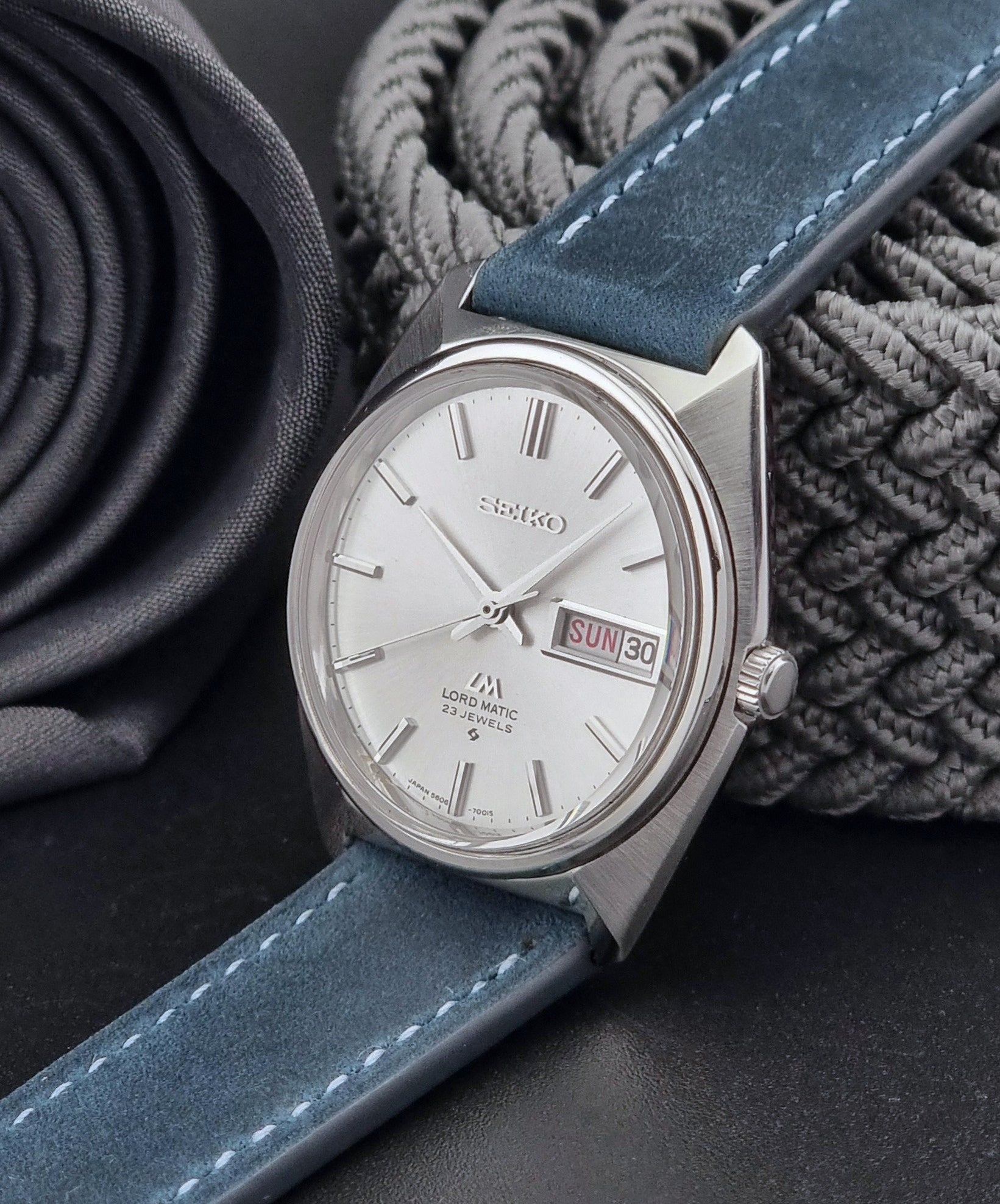 Vintage Seiko Wristwatch LordMatic 5606-7000 with blue strap on a textured gray background crown view