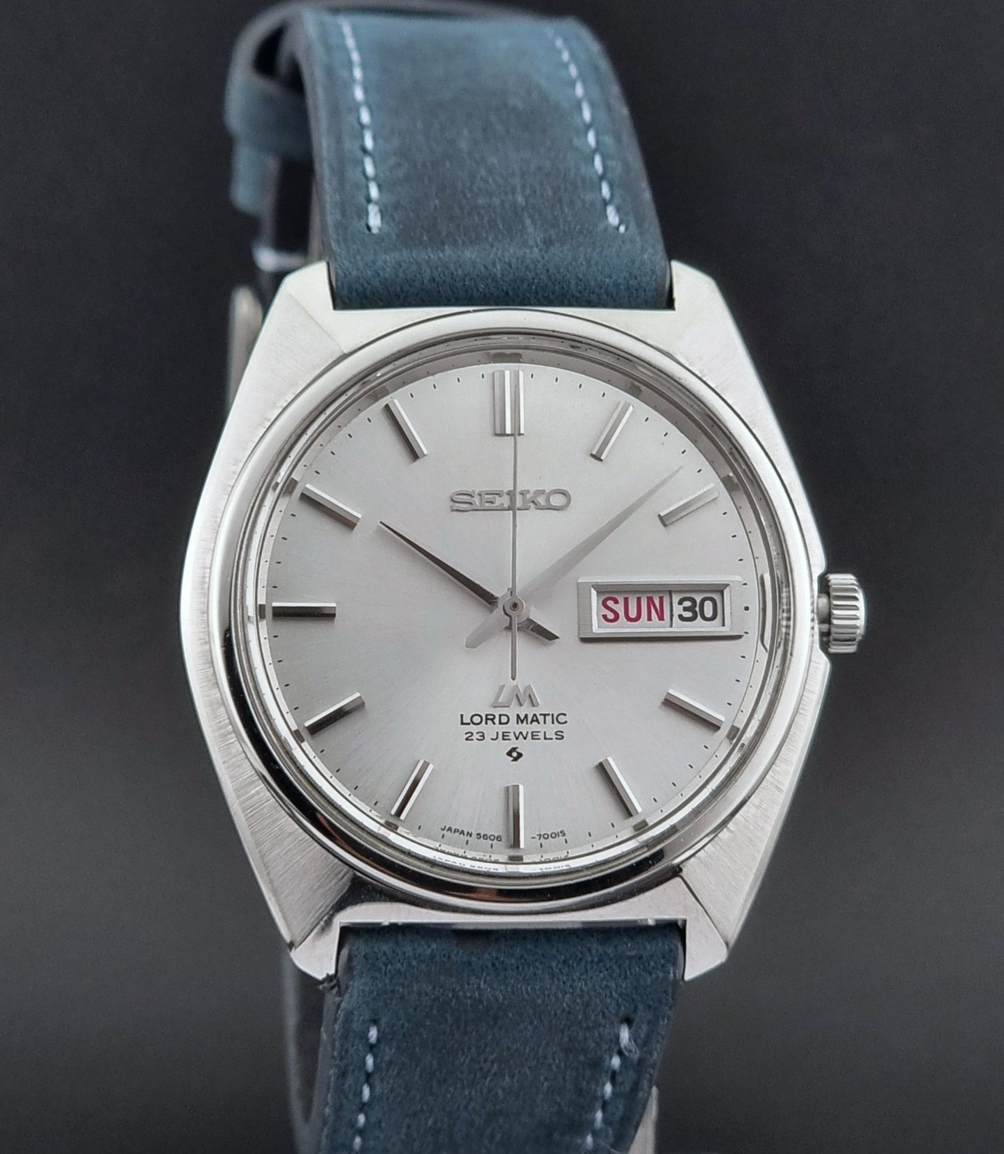 Vintage Seiko Wristwatch LordMatic 5606-7000 with blue strap on a textured gray background front view