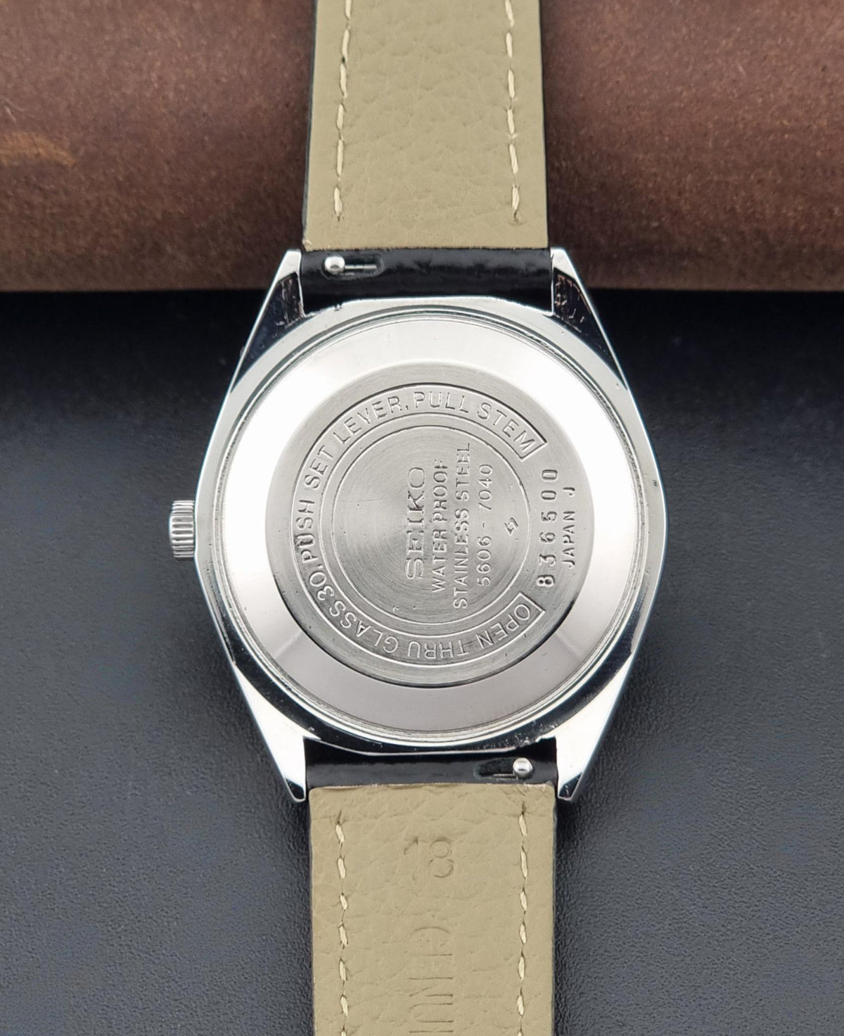 Close-up of the back of A Vintage Seiko watch LordMatic 5606-7040 with a silver metal case, resting on a dark surface with a brown textured background.