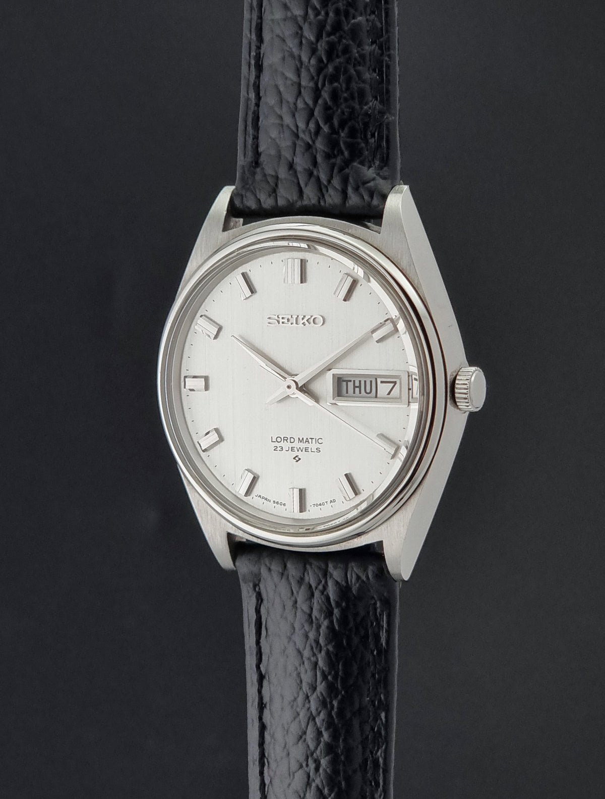 A Vintage Seiko watch LordMatic 5606-7040 with a silver-toned oval case, white dial, and a textured black leather strap, shown against a dark gray background.