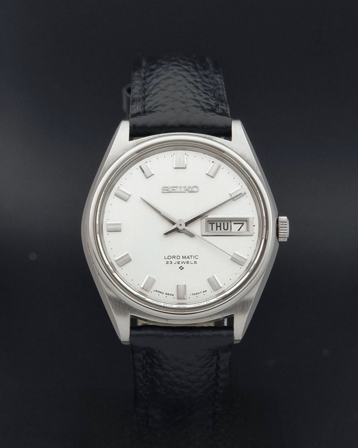 A Vintage Seiko watch LordMatic 5606-7040 with a silver stainless steel case, white dial, and textured black leather strap, shown against a dark background.
