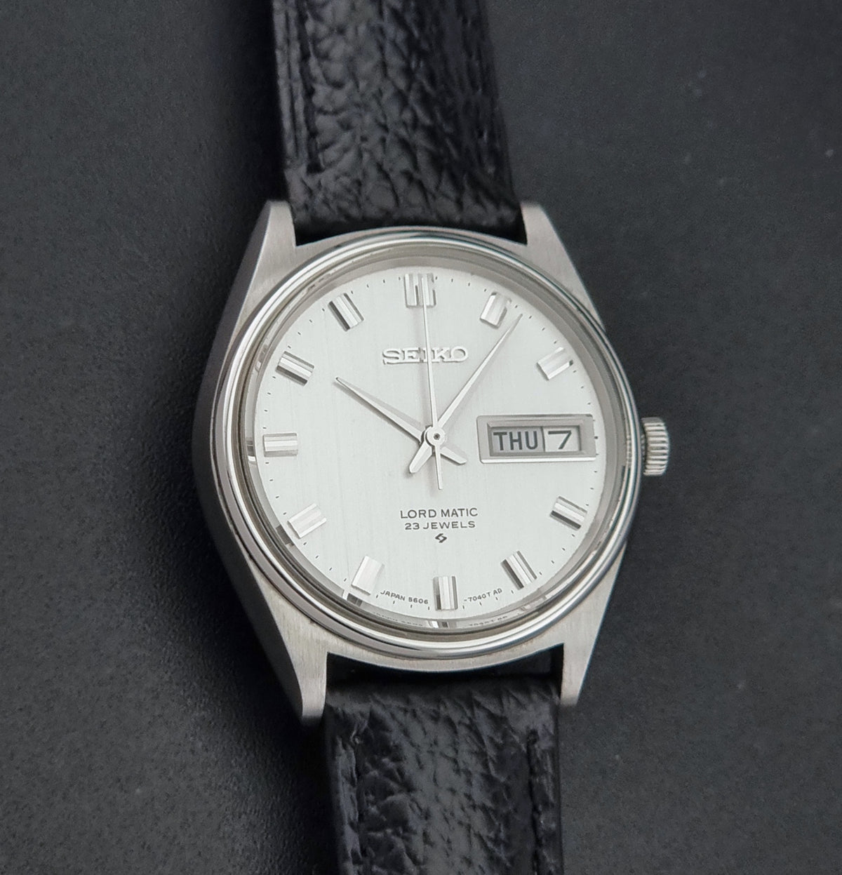 A Vintage Seiko watch LordMatic 5606-7040 with a silver-toned metal case, a white dial with a day-date window, and a textured black leather strap, photographed on a dark gray background.