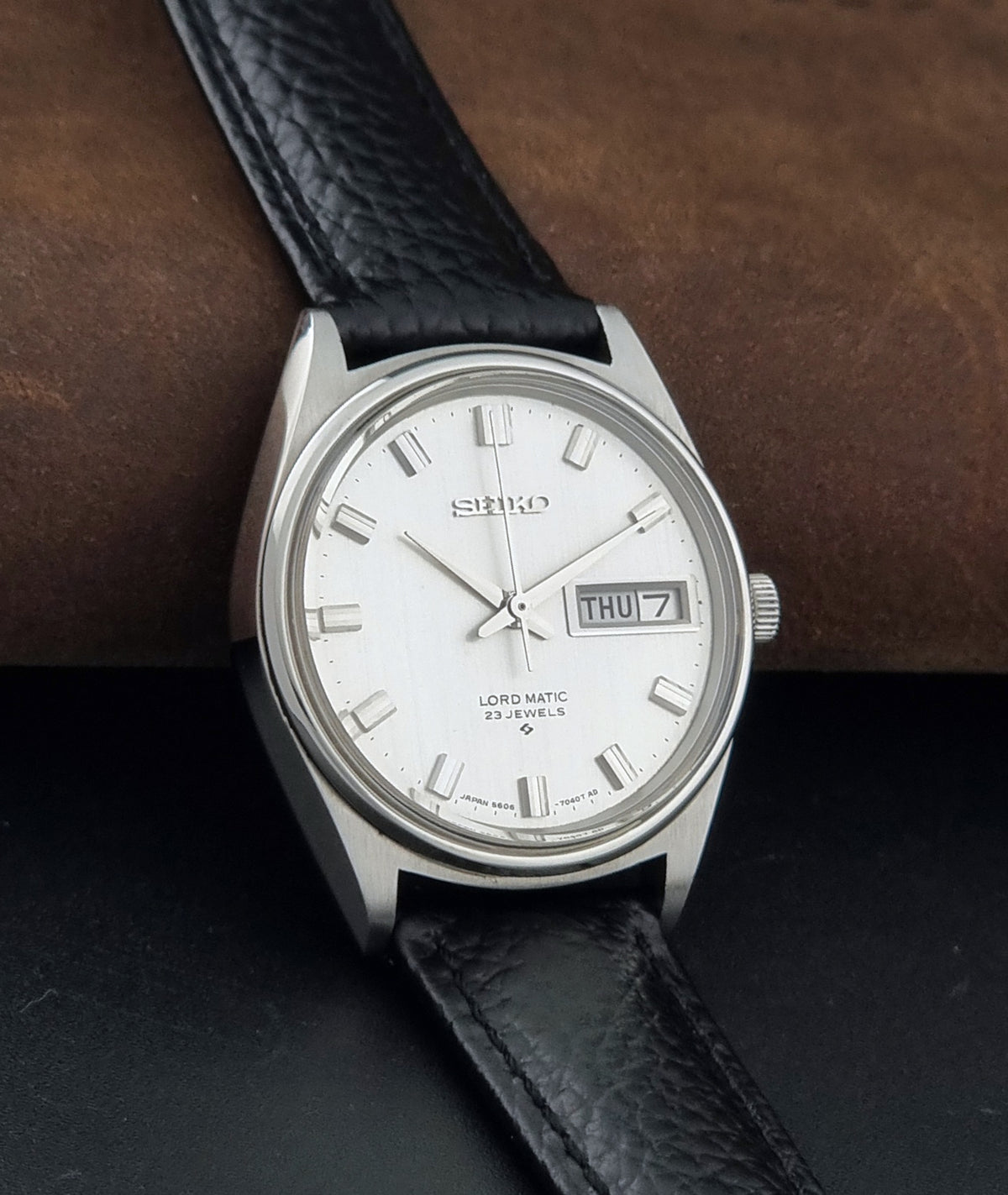 A Vintage Seiko watch LordMatic 5606-7040 with a silver stainless steel case, white dial, and black leather strap, resting on a dark surface with a brown textured background.