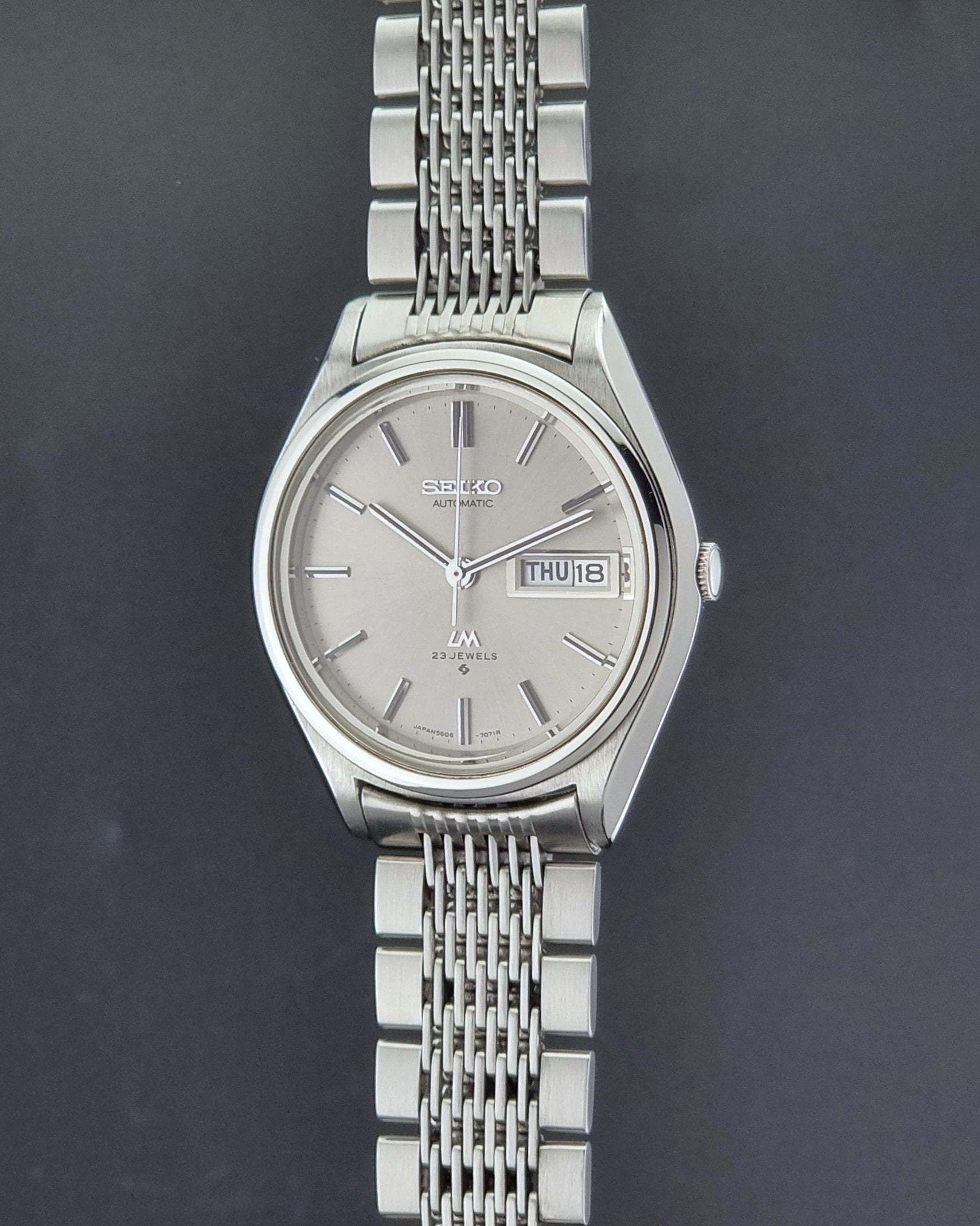 Vintage Seiko 5606-7071 LordMatic wristwatch with a metallic band on a dark gray background crown profile view