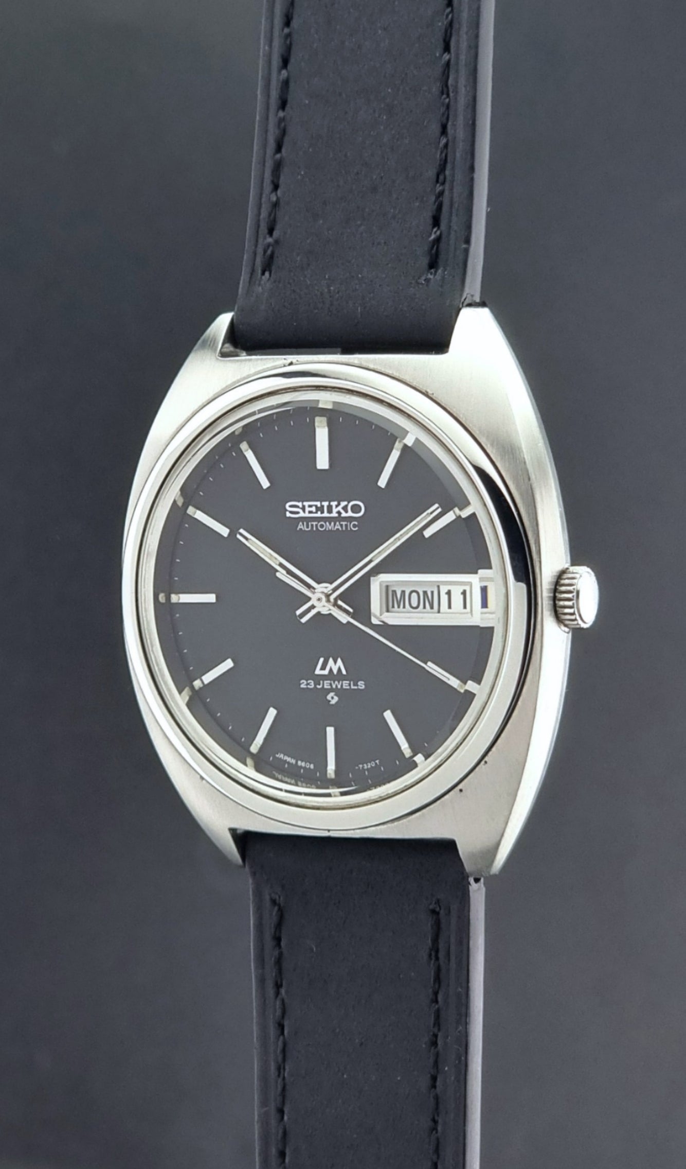 Vintage Seiko Lord Matic 5606-7150 Watch with black strap and gray dial on a wooden surface crown profile