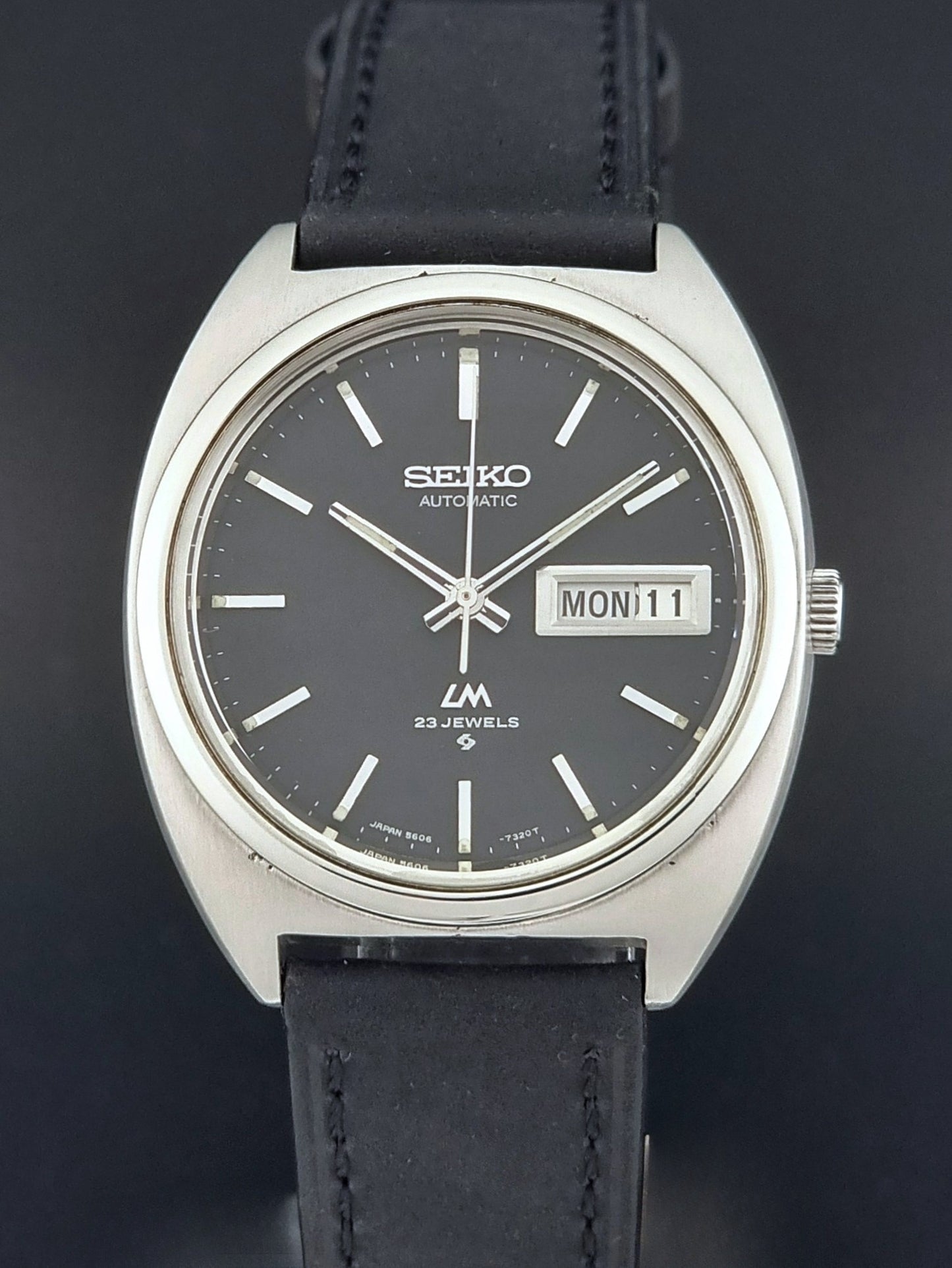 Vintage Seiko Lord Matic 5606-7150 Watch with black strap and gray dial on a wooden surface front view