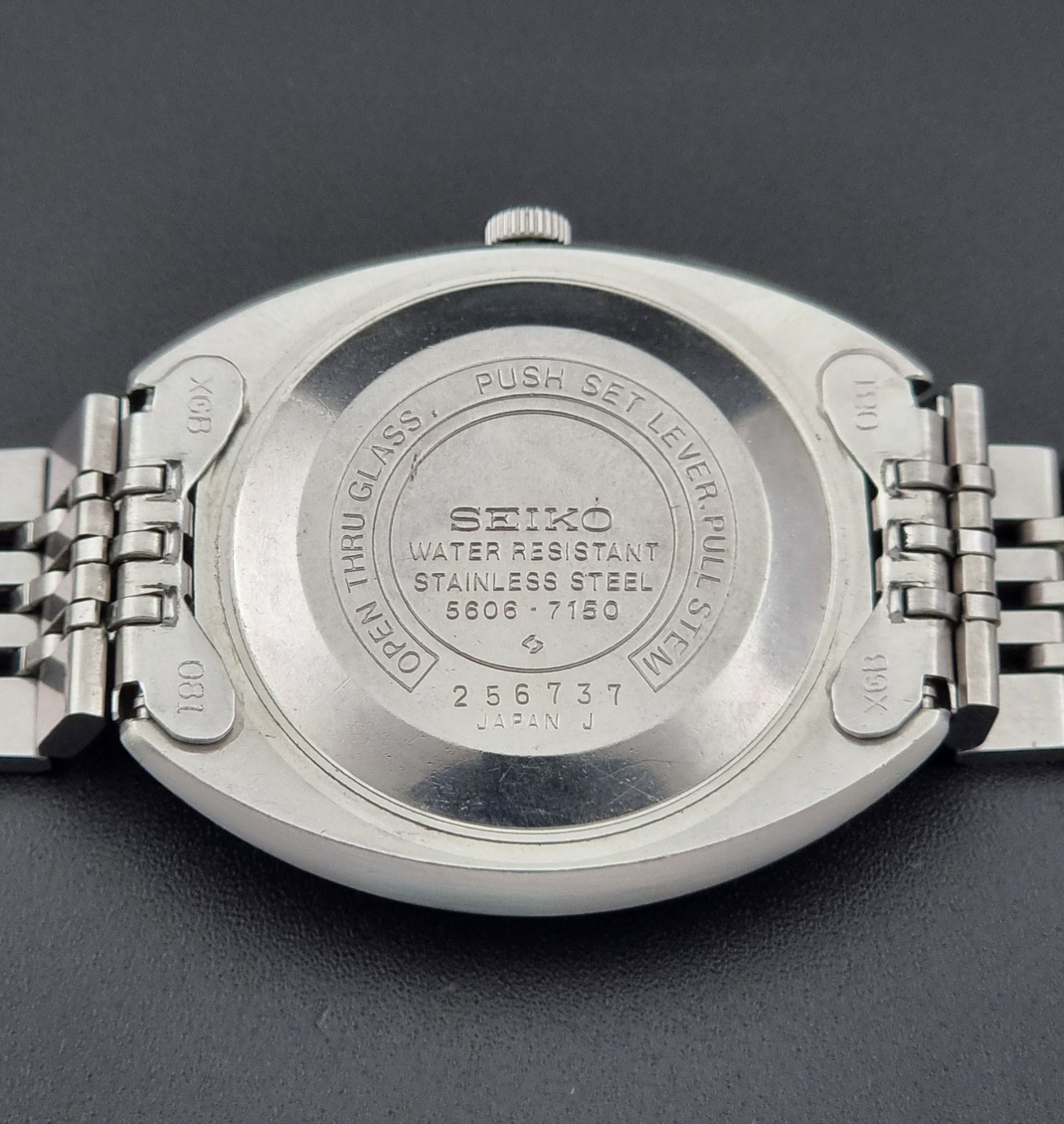 Vintage Seiko Lord Matic 5606-7150 stainless steel wristwatch with a metallic band on a dark gray background close up case back