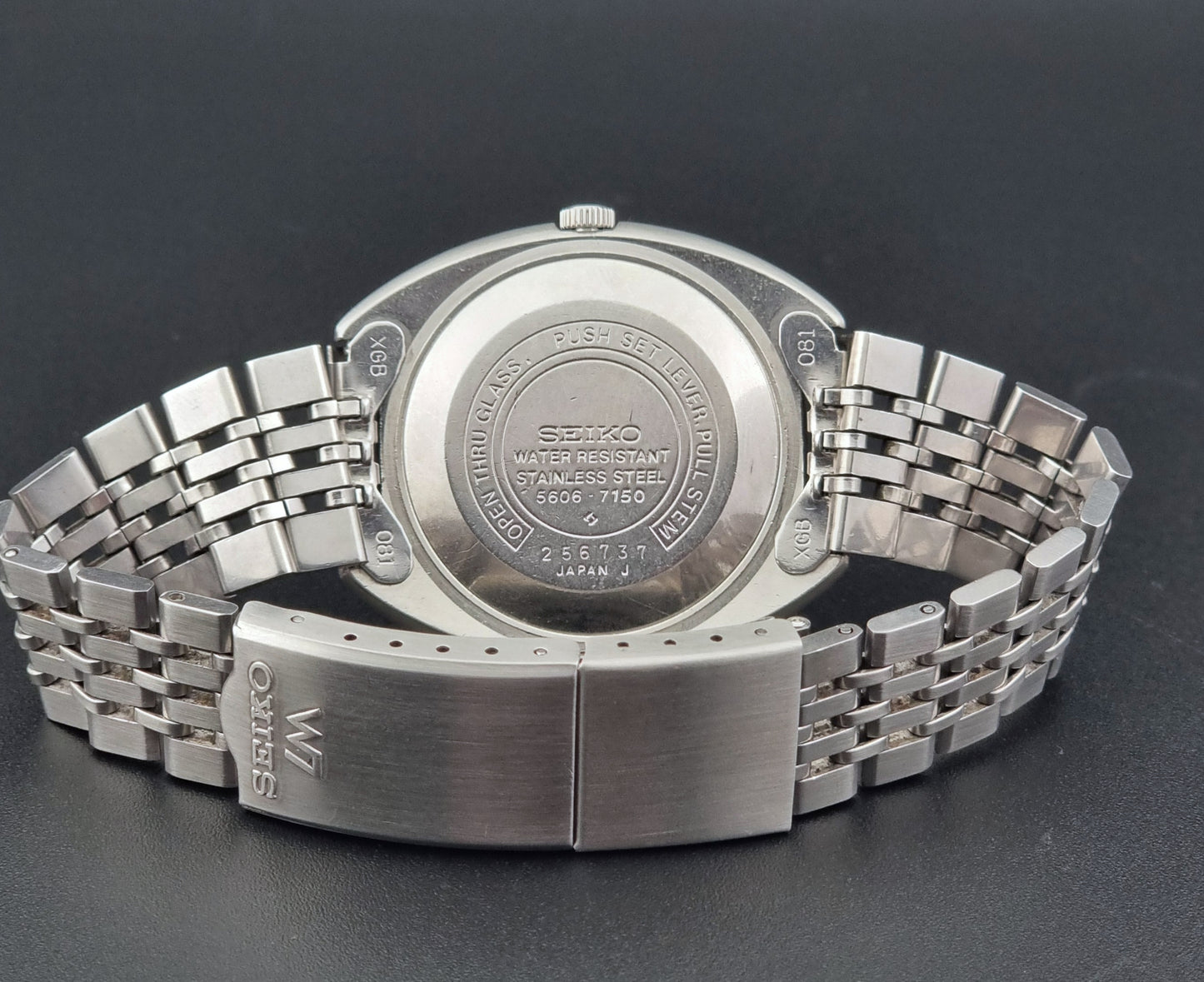 Vintage Seiko Lord Matic 5606-7150 stainless steel wristwatch with a metallic band on a dark gray background case back view