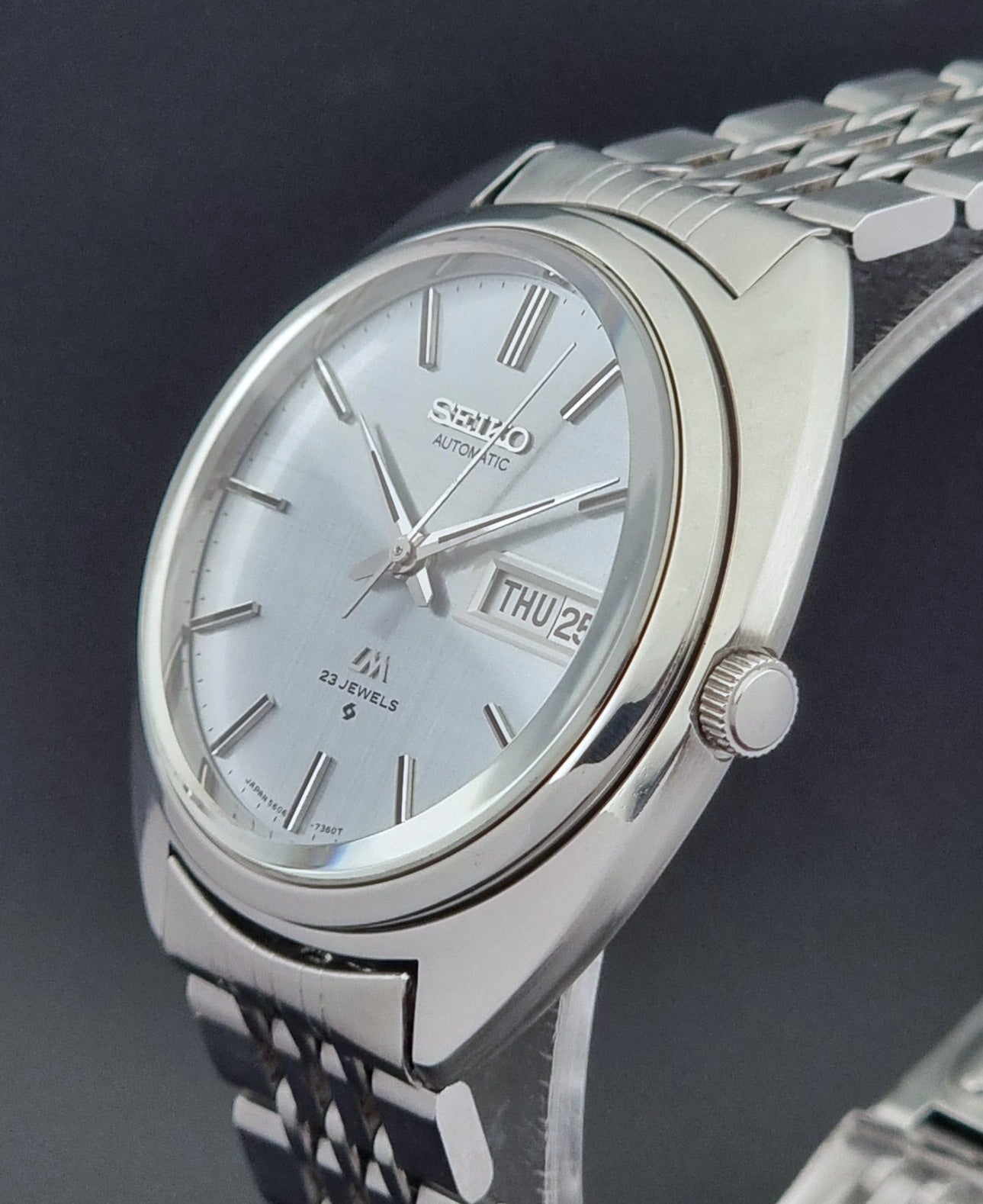 Vintage Seiko Lord Matic 5606-7150 stainless steel wristwatch with a metallic band on a dark gray background crown