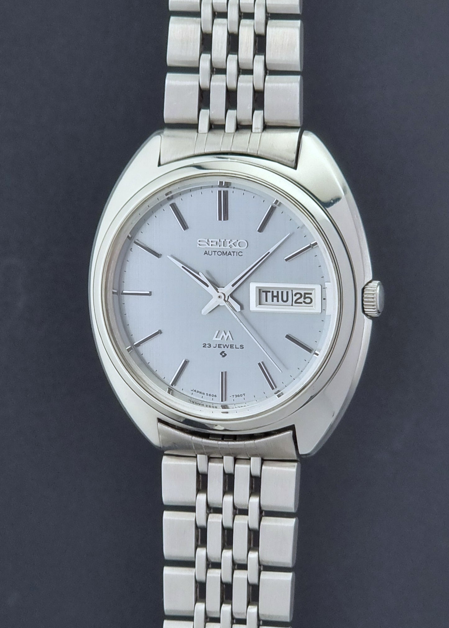 Vintage Seiko Lord Matic 5606-7150 stainless steel wristwatch with a metallic band on a dark gray background crown profile