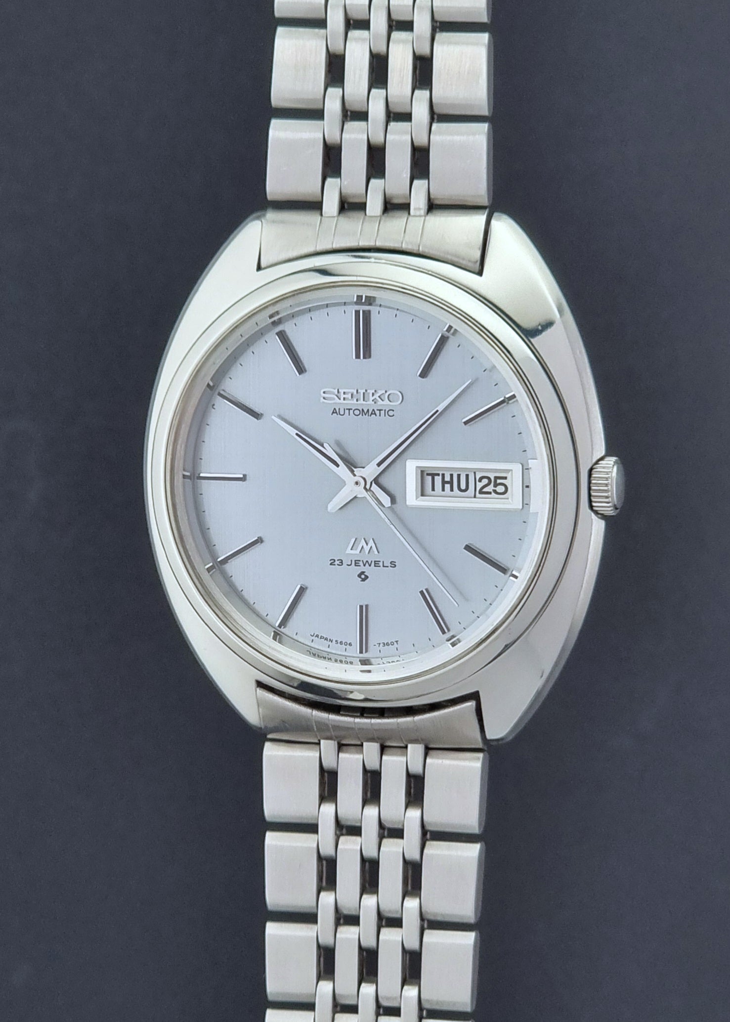 Vintage Seiko Lord Matic 5606-7150 stainless steel wristwatch with a metallic band on a dark gray background crown profile