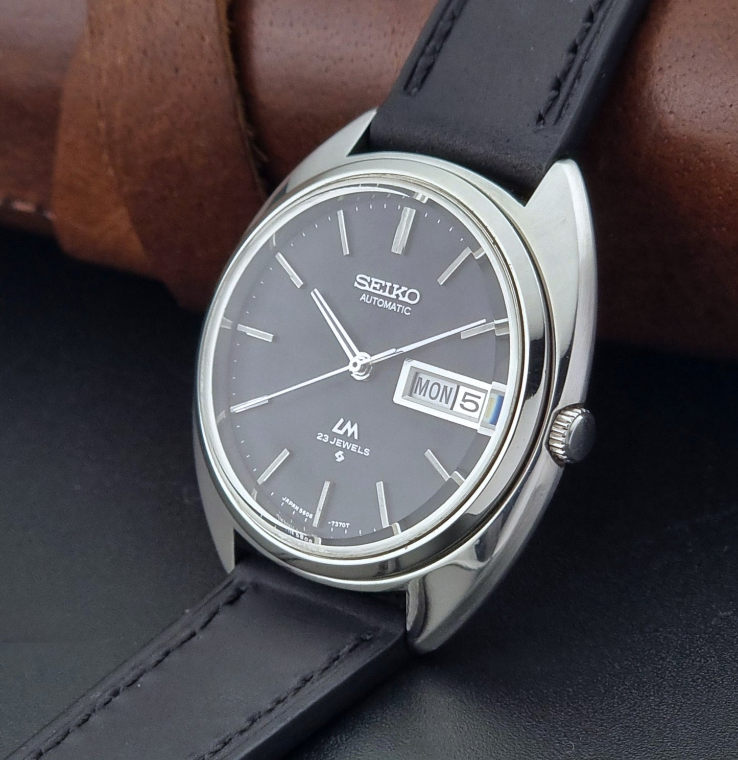 Vintage Seiko watch LordMatic 5606-7150 with black leather strap on a brown leather surface crown side view